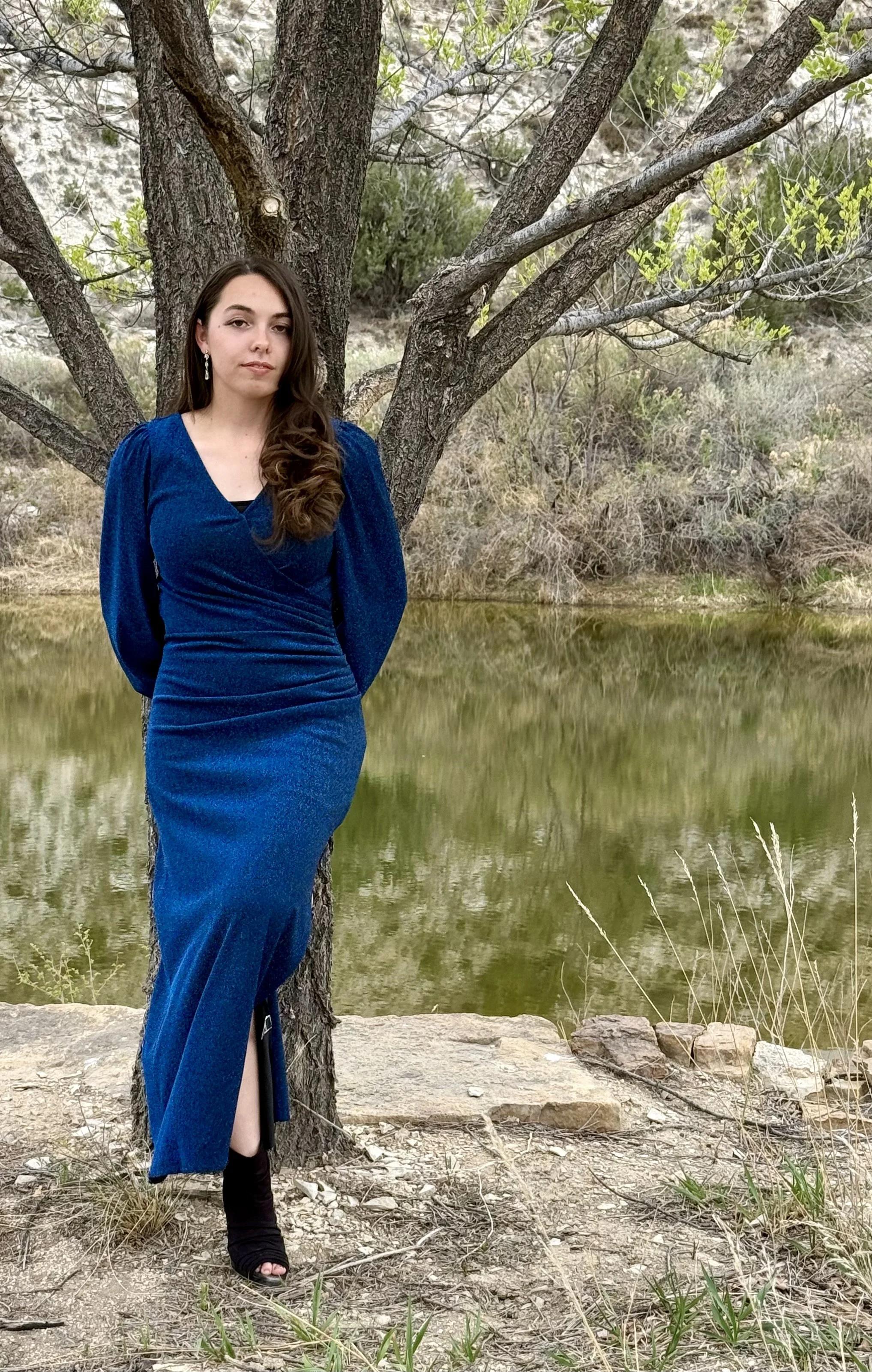 Engaged to be married woman stands by ceremony Elm Tree by the pond at the outdoor waterside wedding ceremony site at Lost Pond wedding venue. Lost Pond is known for its natural water features and beautiful wildflowers and mountain views near Pueblo