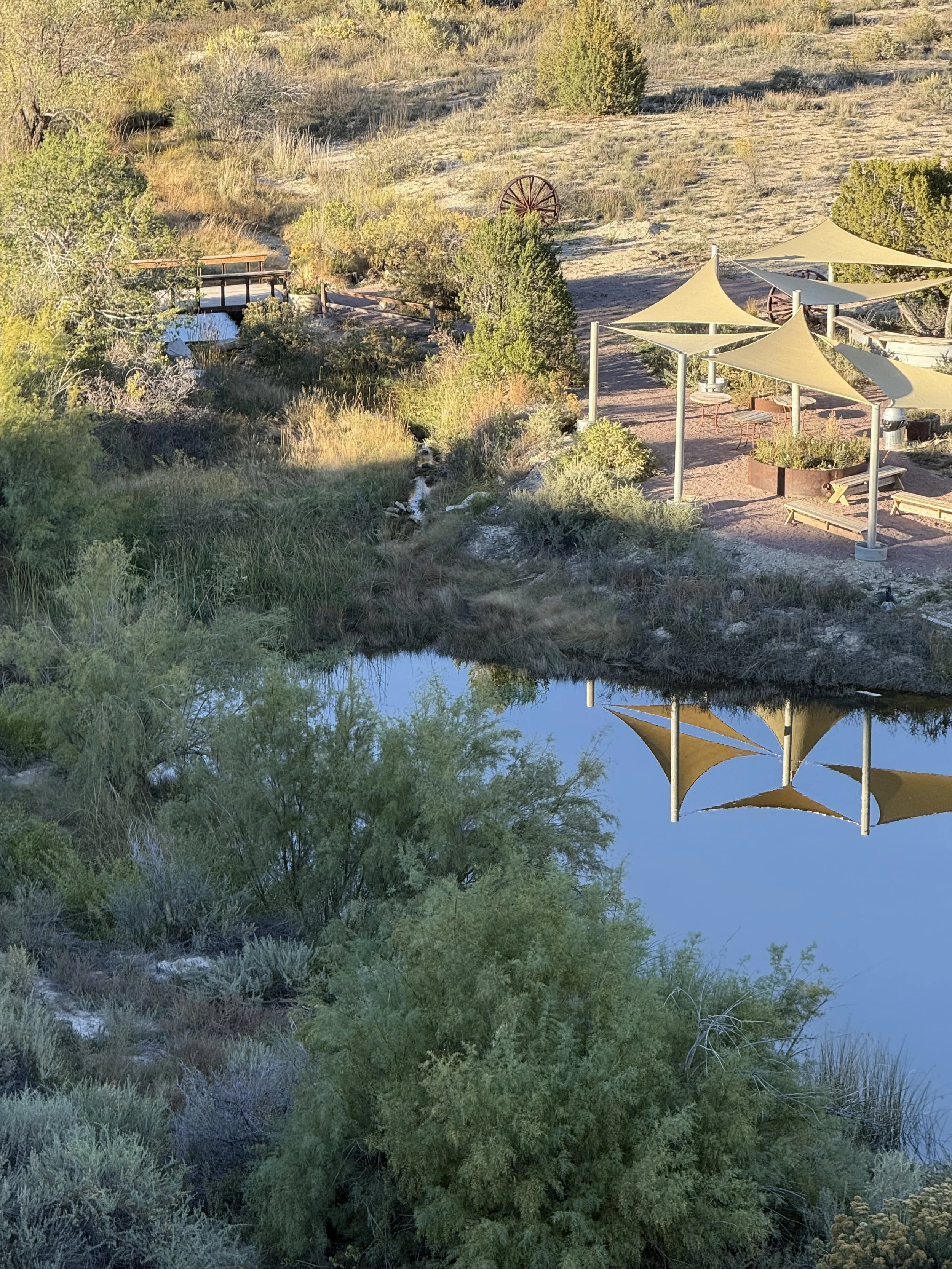 Rustic bridge over the stream with the shaded cocktail hour area beside the pond at Lost Pond wedding venue located between Colorado Springs, and Canon City, Colorado.