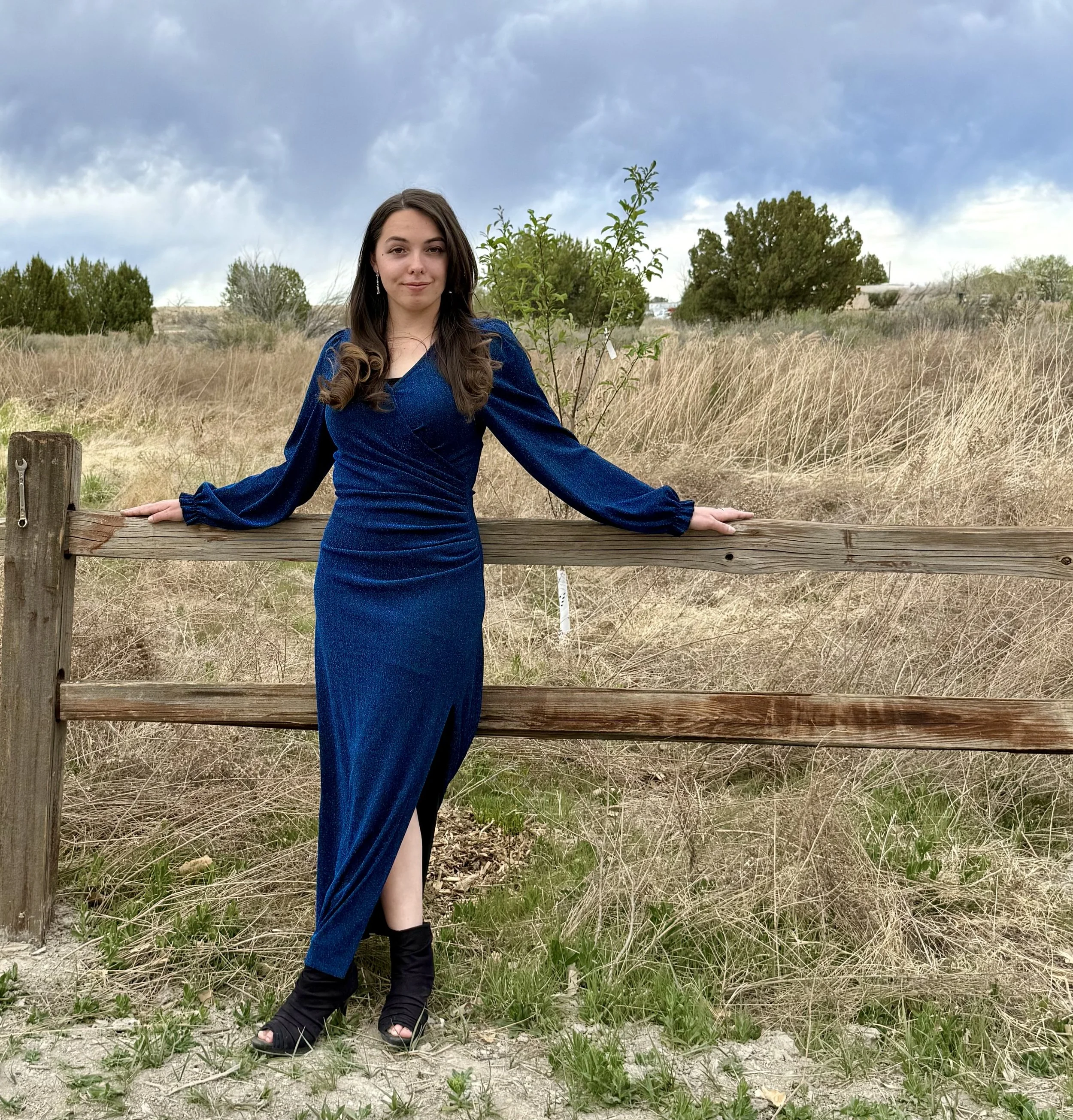 A young woman poses outdoors with one foot on a wooden fence, surrounded by tall grass and trees near a stream at Lost Pond wedding venue. A wedding venue with natural water features and wildflowers and mountain views,  Near Pueblo, Colorado