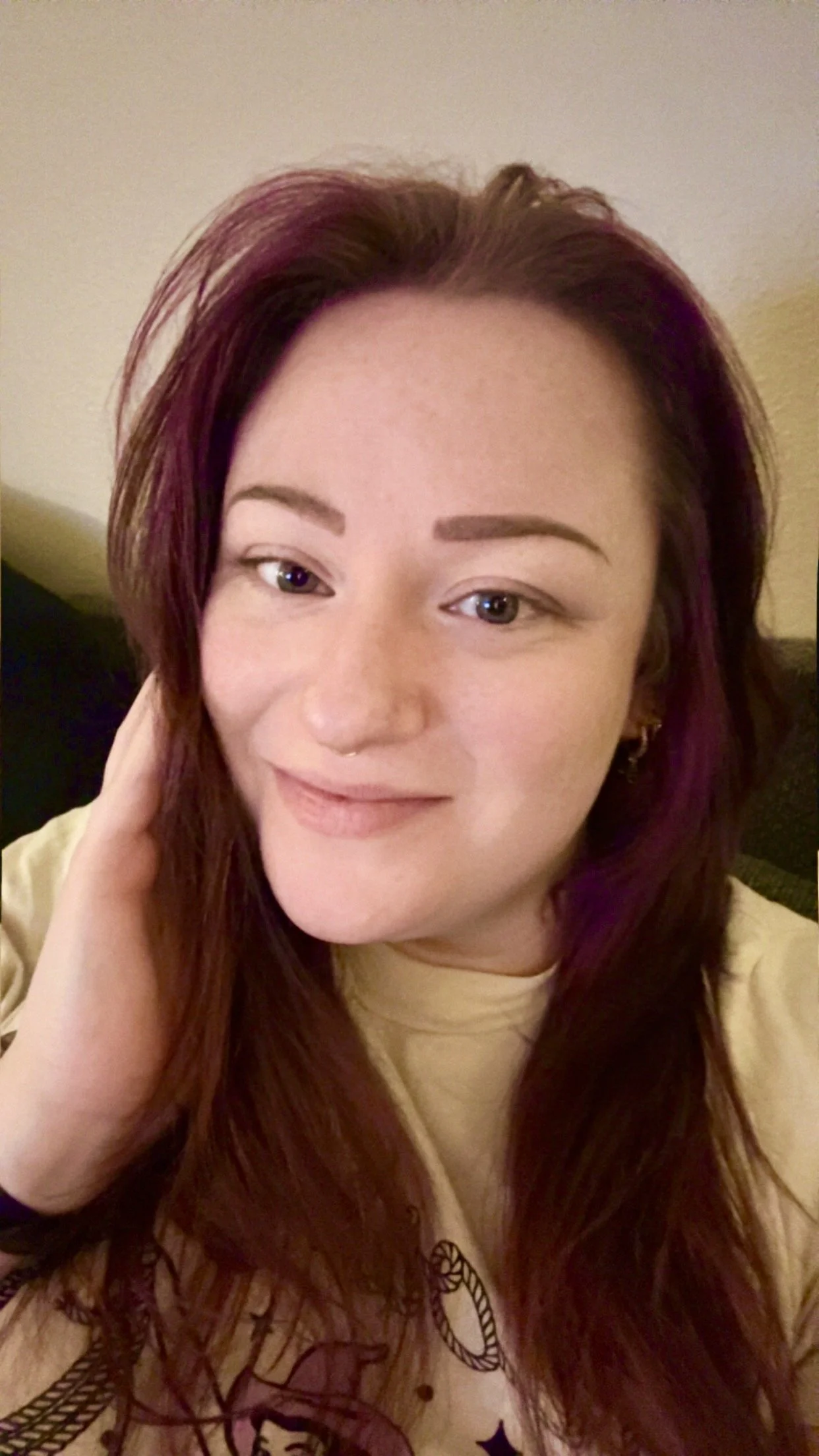 A woman with long reddish-brown hair, blue eyes, light skin, wearing a light-colored shirt and earrings, smiling slightly and touching her hair.