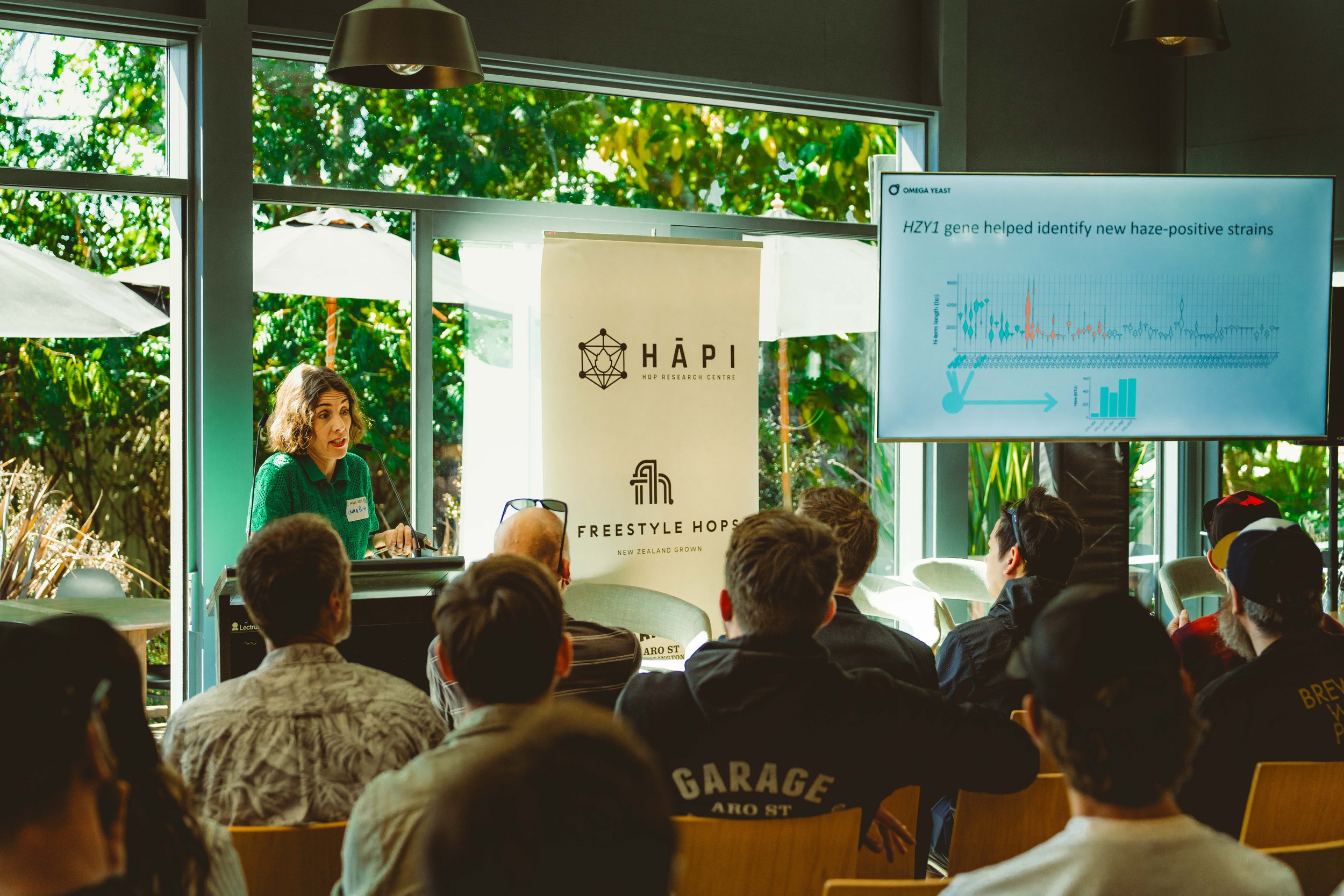 A woman is giving a presentation to an audience in a room with large windows and green outdoor scenery. There is a large screen displaying a graph and text, with banners for HĀPI and FREESTYLE HOPS.