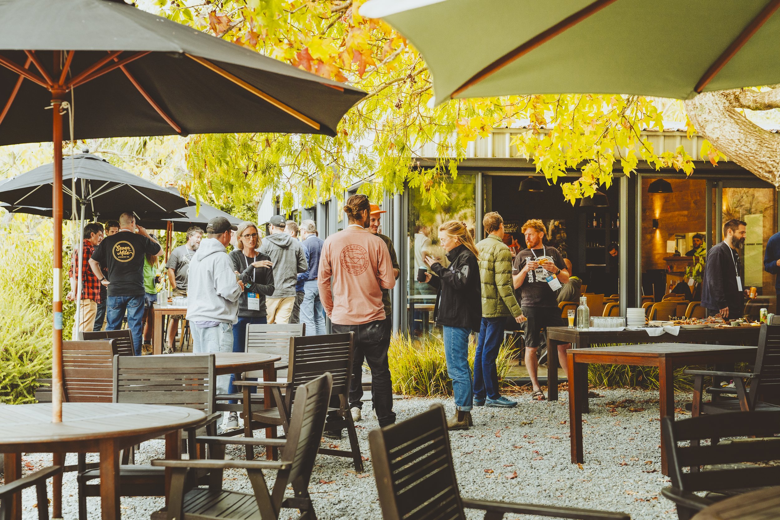 People gathered outdoors at a gathering or event, standing and socializing under umbrellas surrounded by autumn trees and tables with chairs, with some inside a building visible through a large glass window.