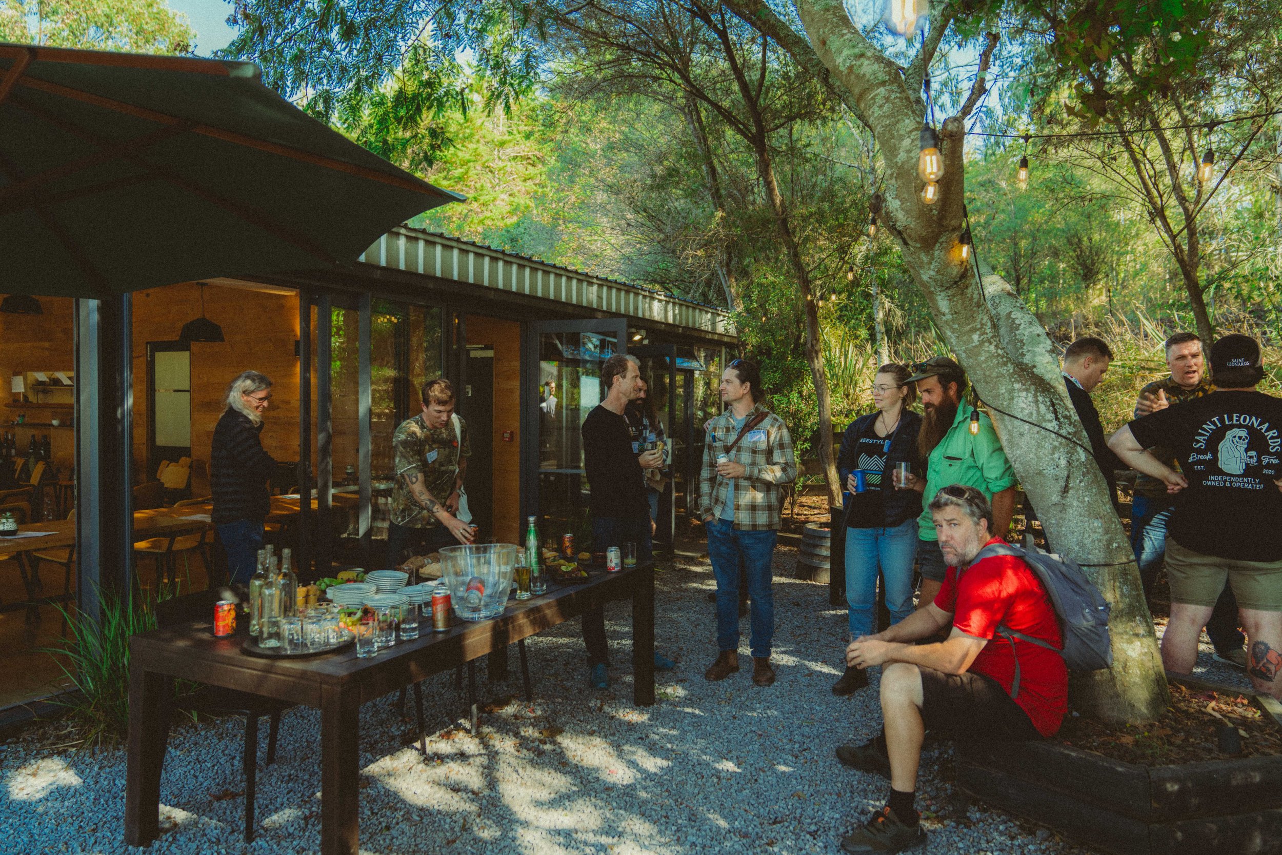 People gathering outdoors near a wooden building with a table of drinks and snacks, surrounded by trees with string lights hanging, on a sunny day.