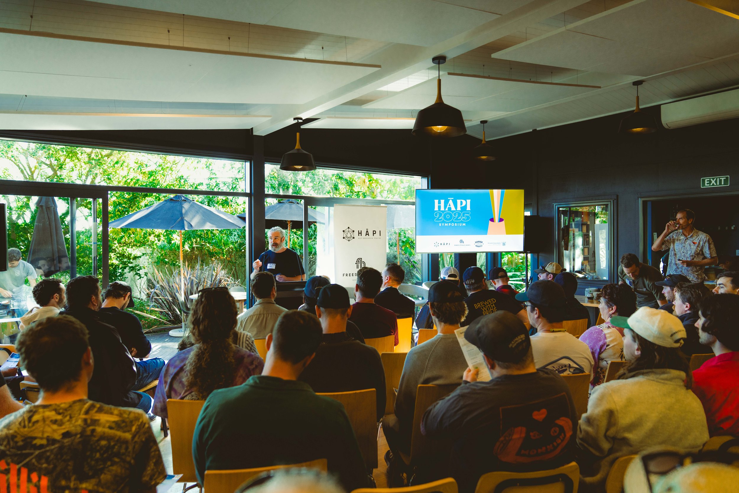 Audience attending a symposium presentation, with a speaker at a podium, a large screen displaying 'HĀPI 2025 Symposium,' and banners, in a room with large windows showing outdoor umbrellas and greenery.