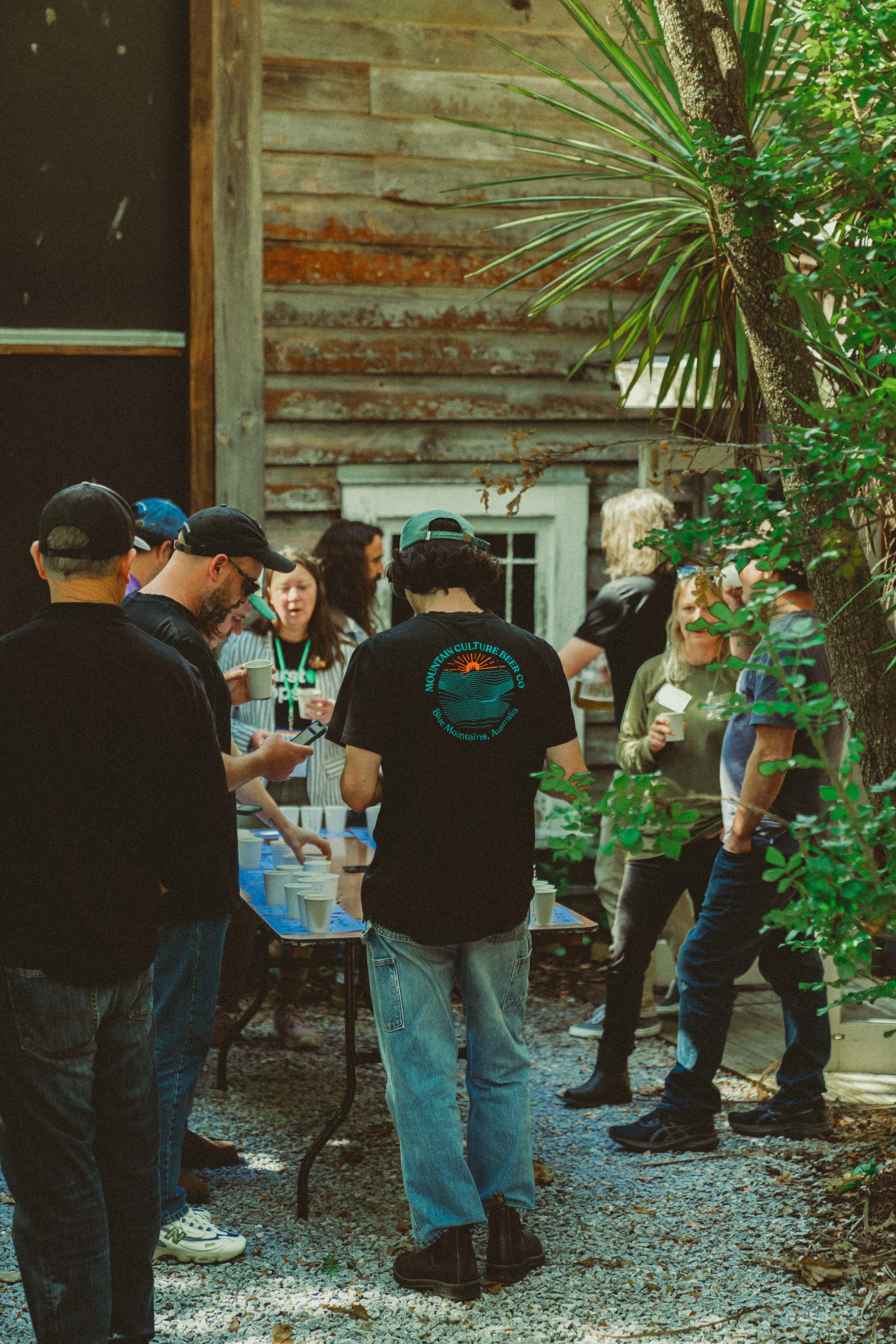Group of people gathered outdoors around a table with cups, socializing near a rustic wooden building and trees.