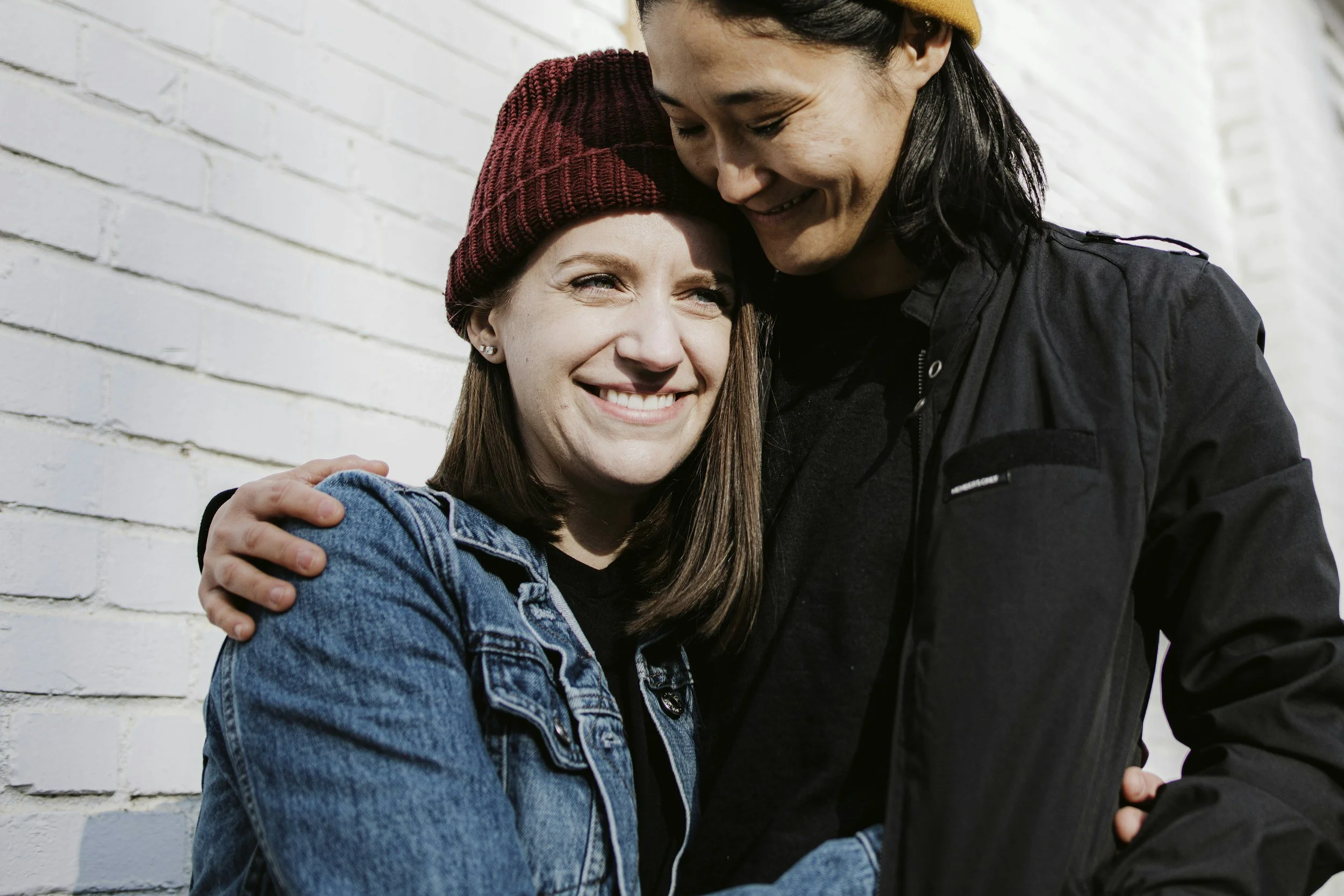 A smiling woman wearing a denim jacket and maroon beanie hugging a man with black hair and a black jacket, standing close against a white brick wall.