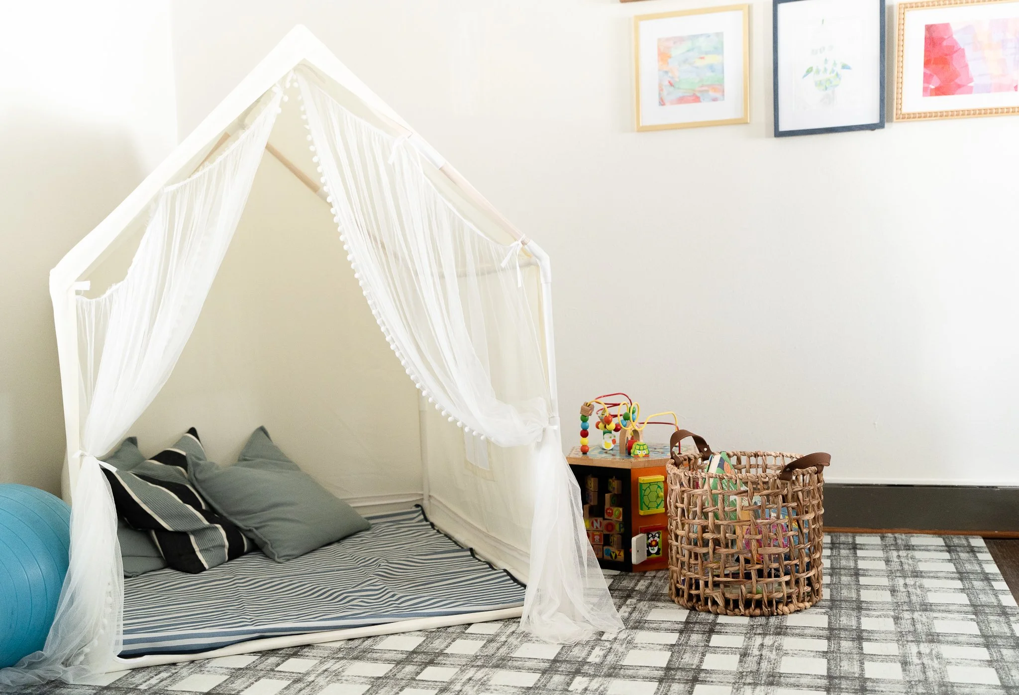 A children's play area with a white canopy tent, gray pillow set, framed artwork on the wall, a small wooden toy storage with colorful toys, and a woven basket on a checkered rug.