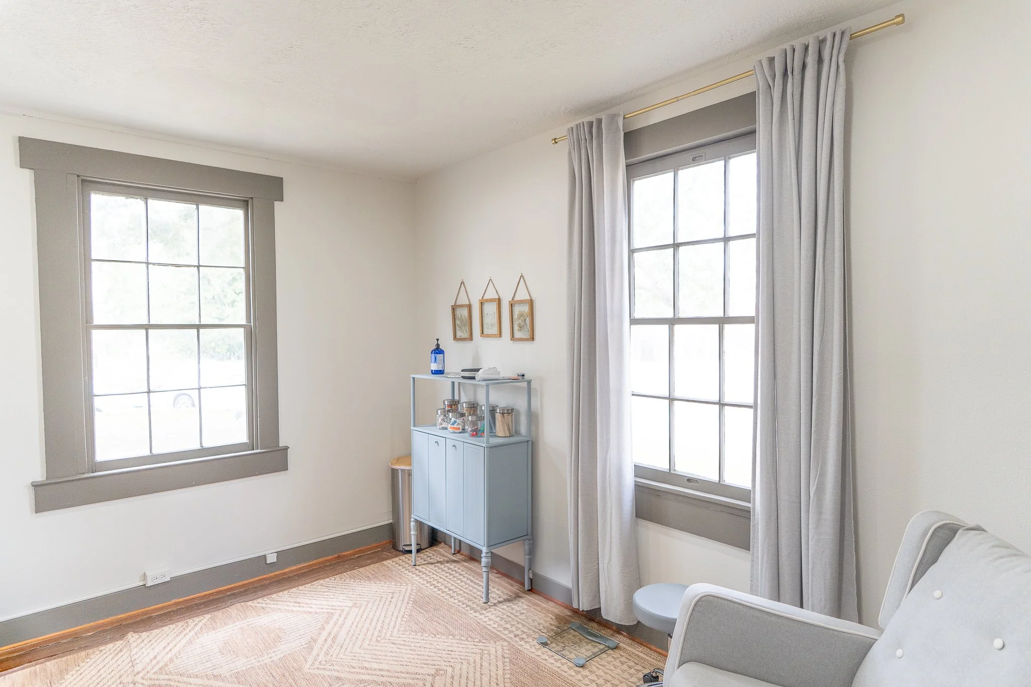 A bright living room corner featuring two large windows with gray trim, sheer curtains, a light blue cabinet with glass jars, a wall-mounted triptych of botanical prints, a white armchair, a digital scale, and a beige carpet on light wooden flooring.