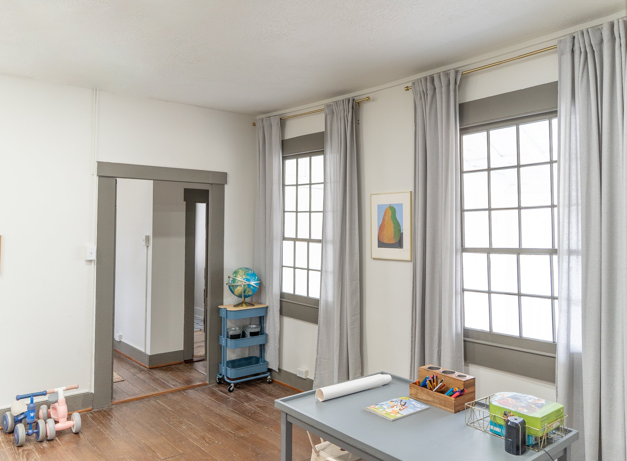 A bright room with large windows covered by gray curtains. There is a table with art supplies, a globe on a small blue cart, and children's ride-on toys on the wooden floor.