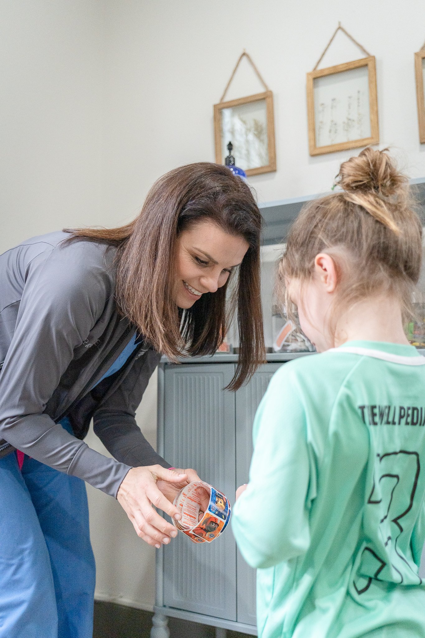 A pediatrician with brown hair smiling as she hands a roll of stickers to a girl with blonde hair in a bun. The girl wears a green shirt with the number 23 on the back. They are in an exam room with a light-colored wall and framed pictures hanging.