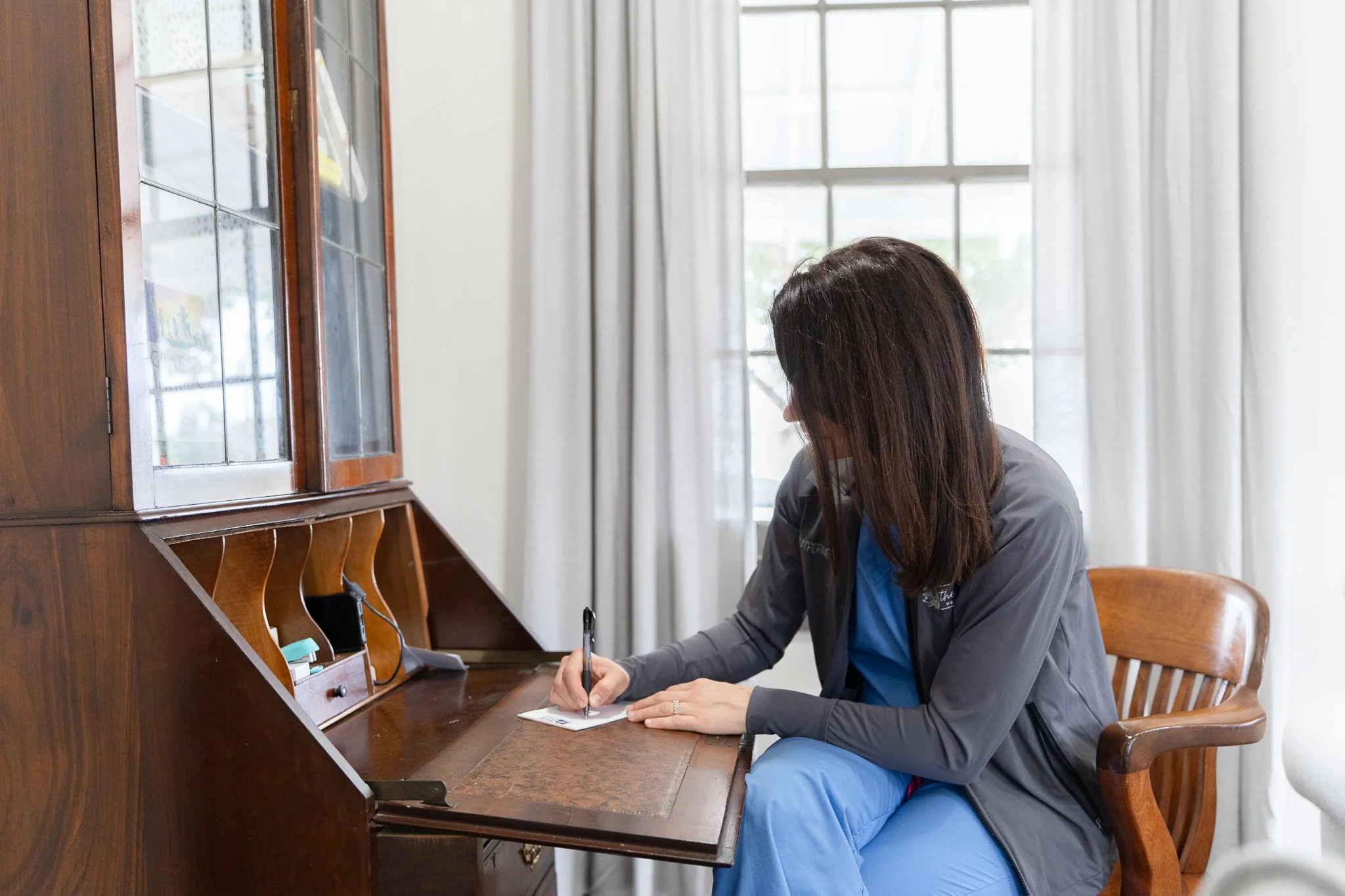 A concierge pediatrician in medical scrubs and a gray jacket sitting at a wooden desk, writing on a notepad in a bright room with large windows and cream curtains.