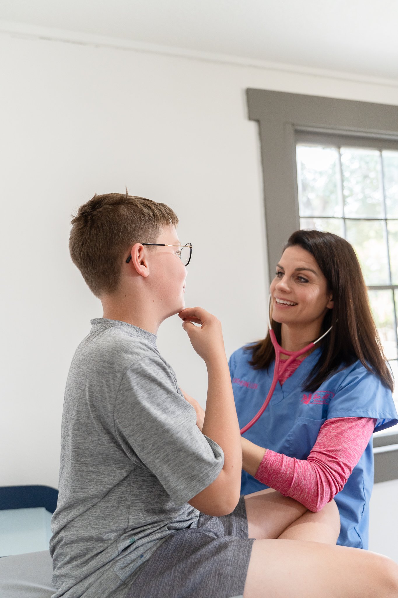 A pediatrician wearing scrubs and a stethoscope listens to a young boy's chest with a smile, sitting on an examination table in a pediatrician's office with a window in the background.