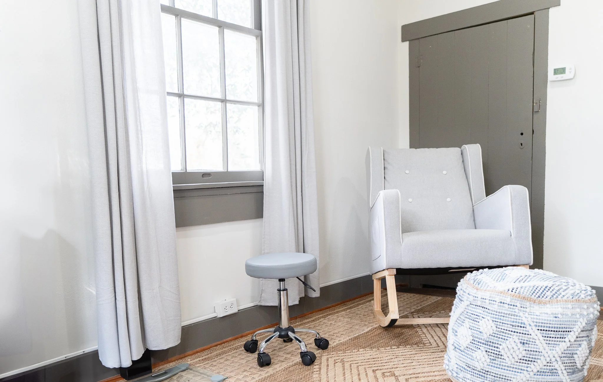 A cozy corner of a pediatric office with a large window with white curtains, a gray rocking armchair, a gray adjustable stool, a woven pouf, and hardwood flooring.