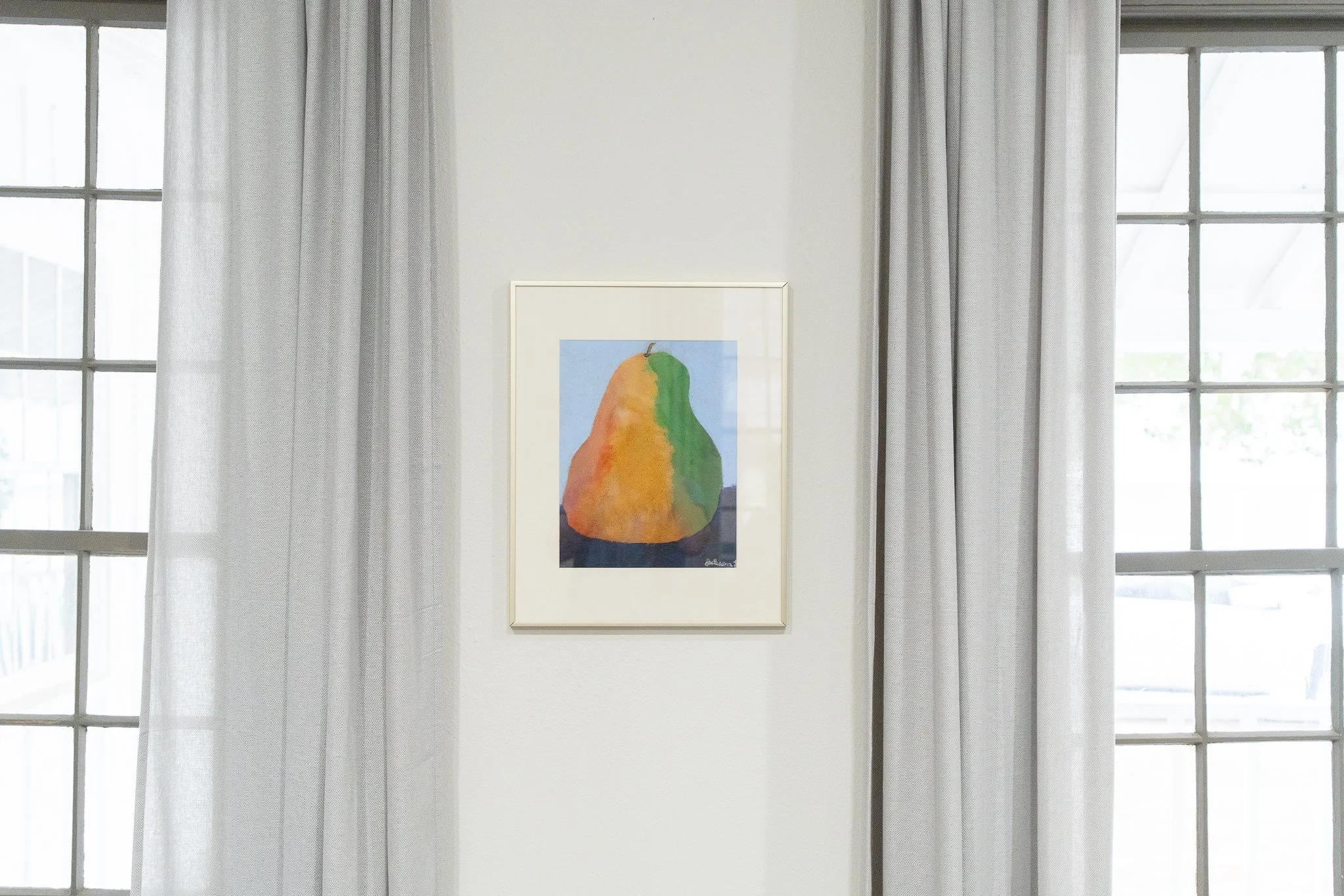 Painting of a pear with orange, green, and yellow colors hanging on a white wall between two windows with white curtains