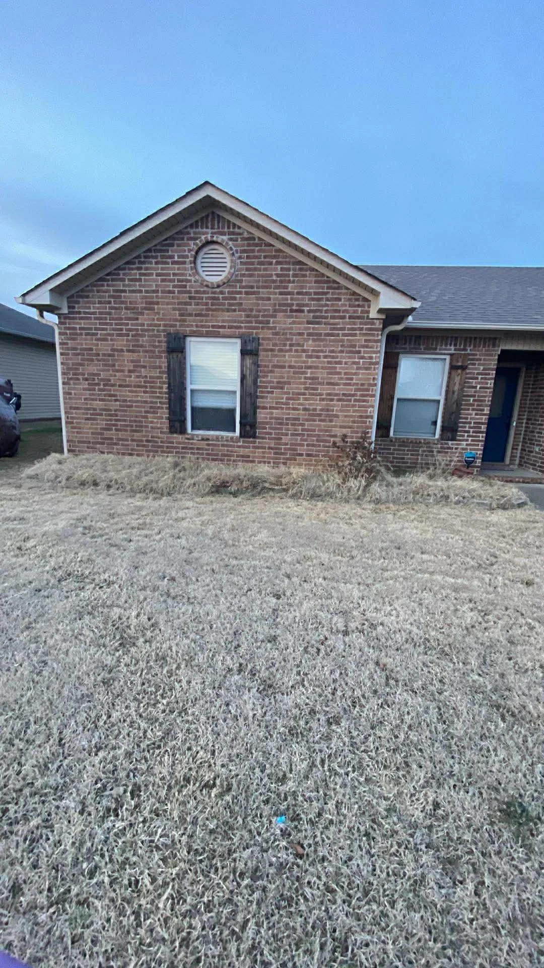A brick house with two windows, one with dark shutters, and a small patch of grass in front; the sky is cloudy and gray.