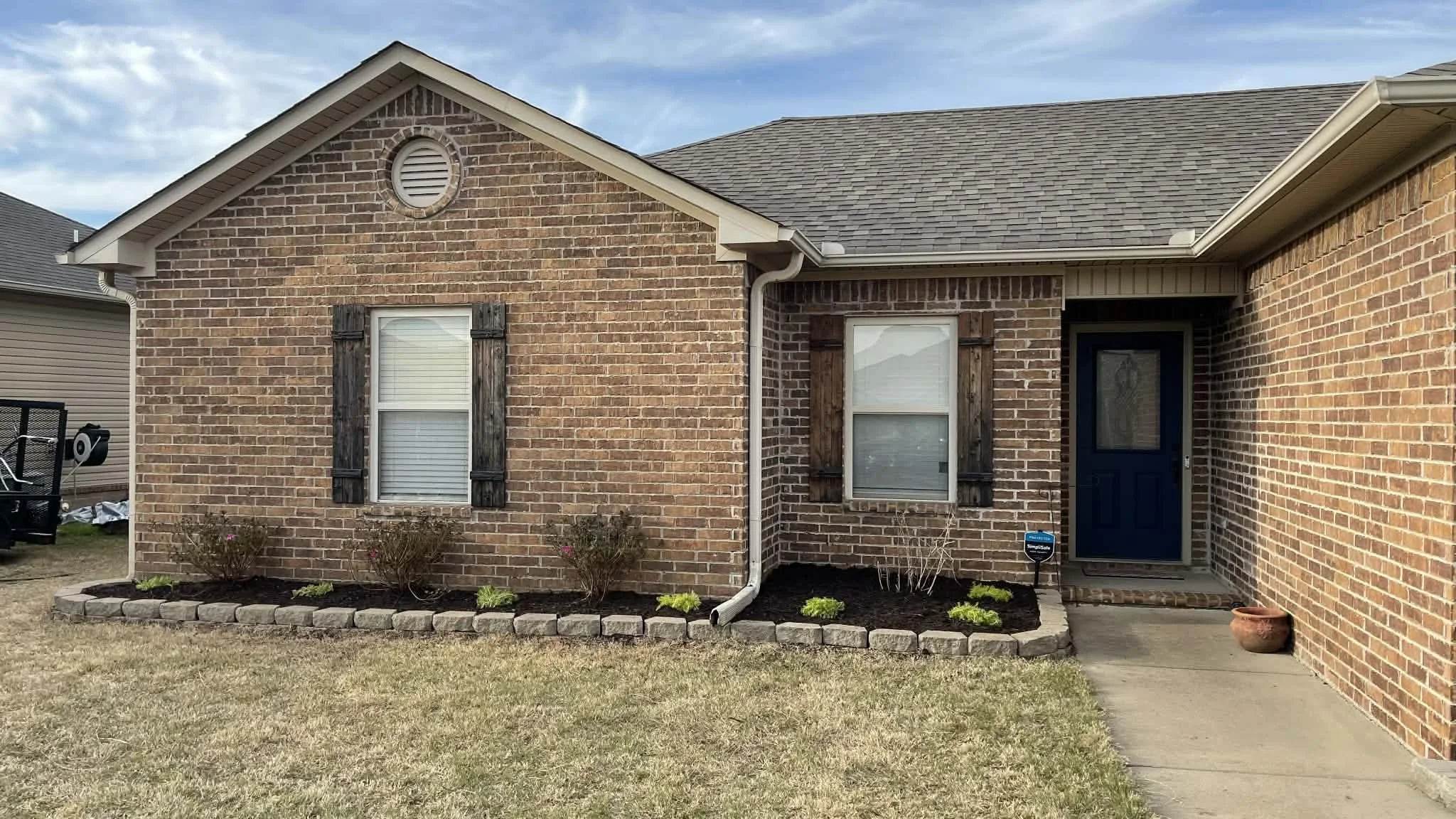 Front view of a brick house with two windows with wooden shutters, a blue front door, a stone-bordered flower bed with small plants, and a concrete walkway leading to the door.