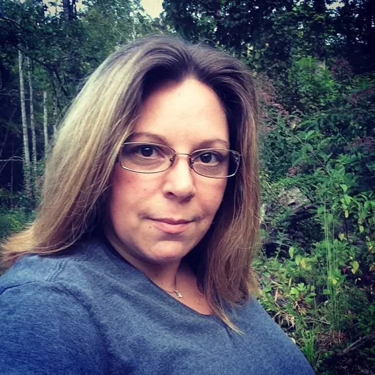 A woman with light brown hair and glasses taking a selfie outdoors with greenery in the background.