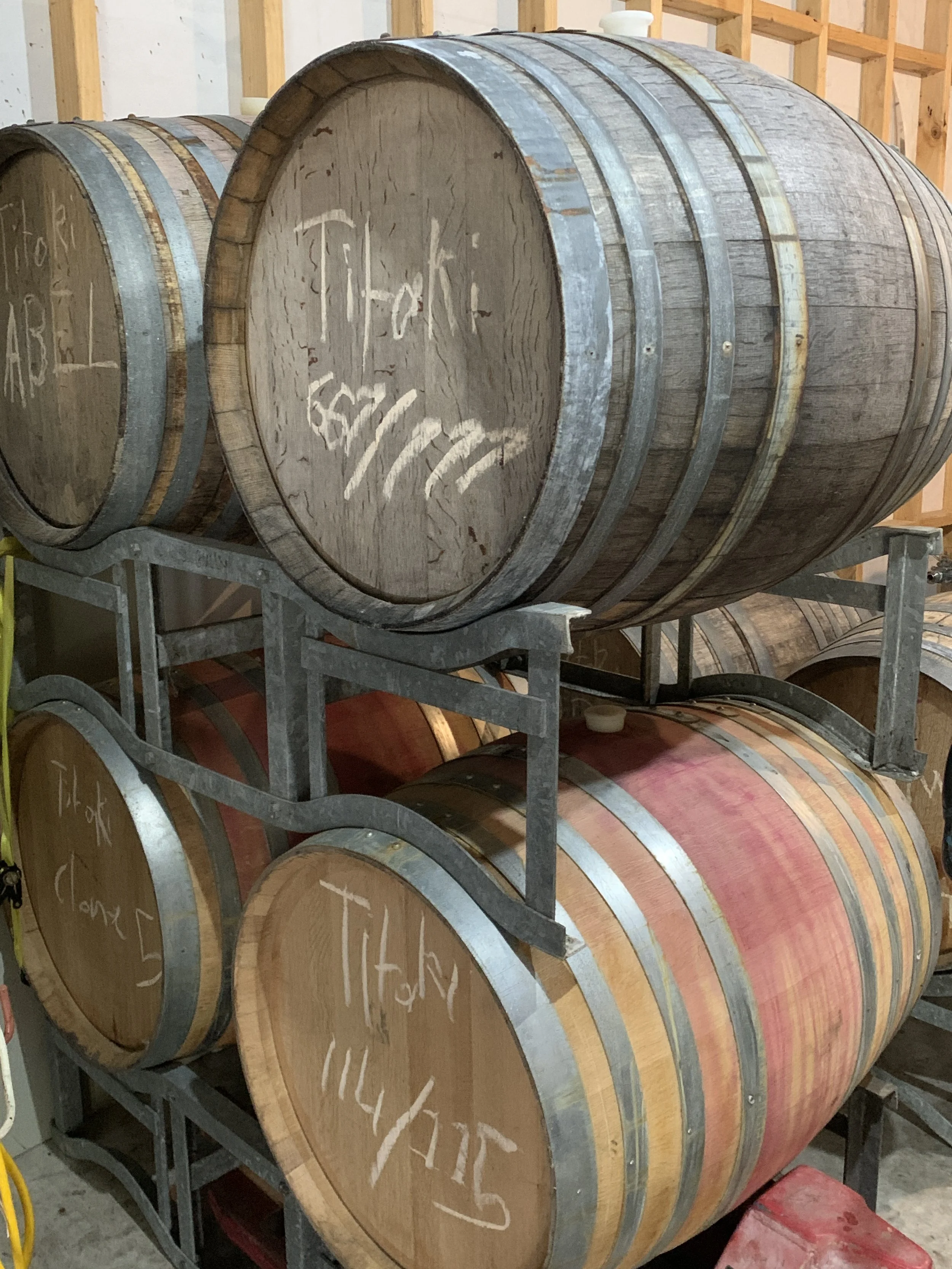 Stacked wooden barrels on metal racks in a cellar, with chalk writing on the ends of the barrels.