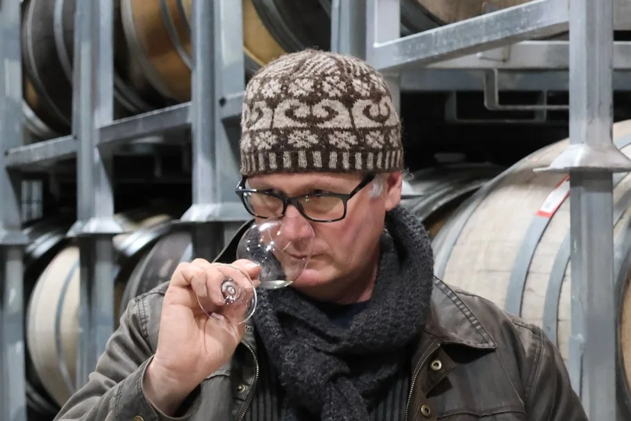 A man with glasses, a knit hat, and a scarf is smelling a glass of wine in a barrel warehouse.