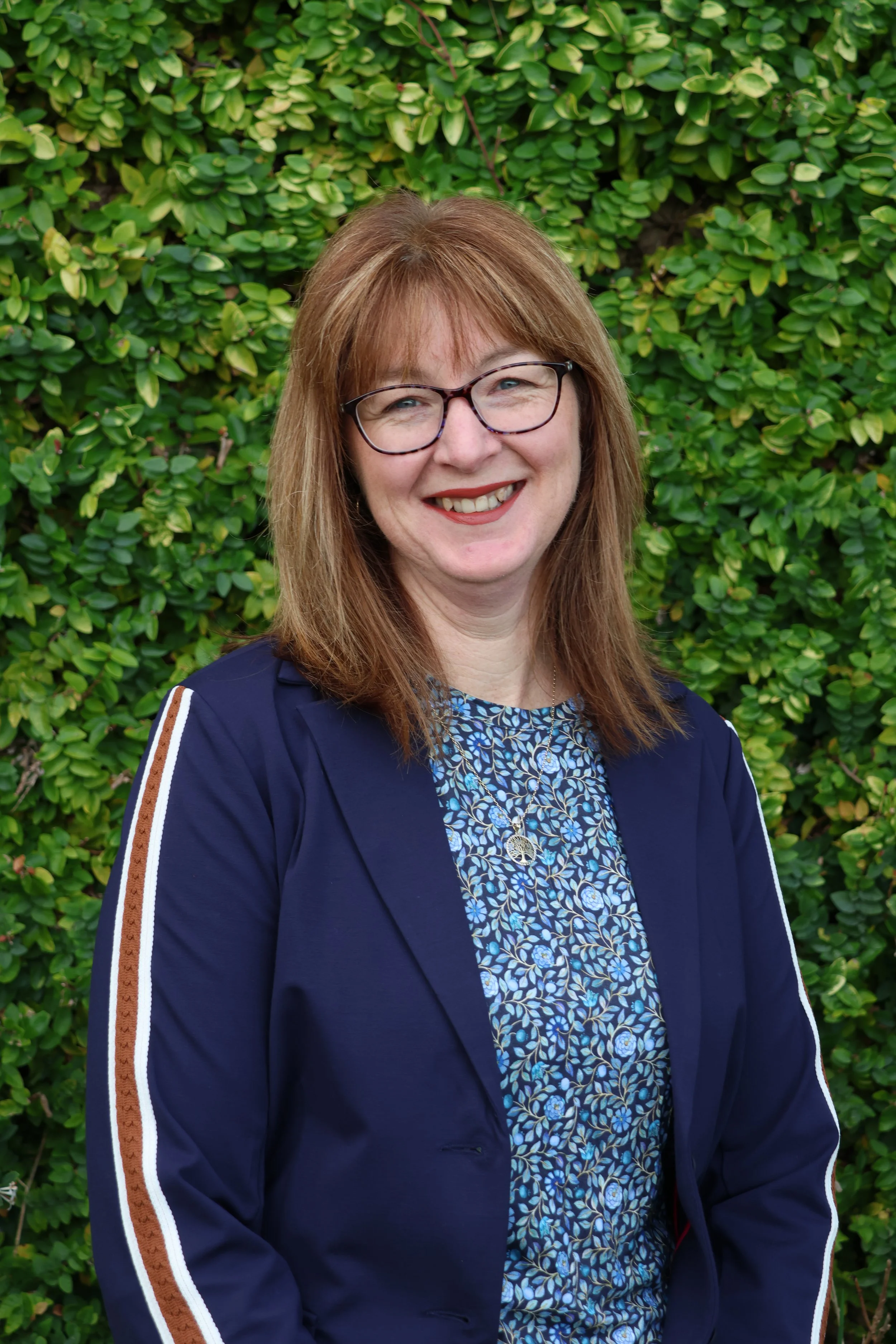 Portrait of a smiling woman with glasses and shoulder-length reddish hair, standing in front of a green leafy background, wearing a blue floral blouse and a navy blazer with striped embellishments.