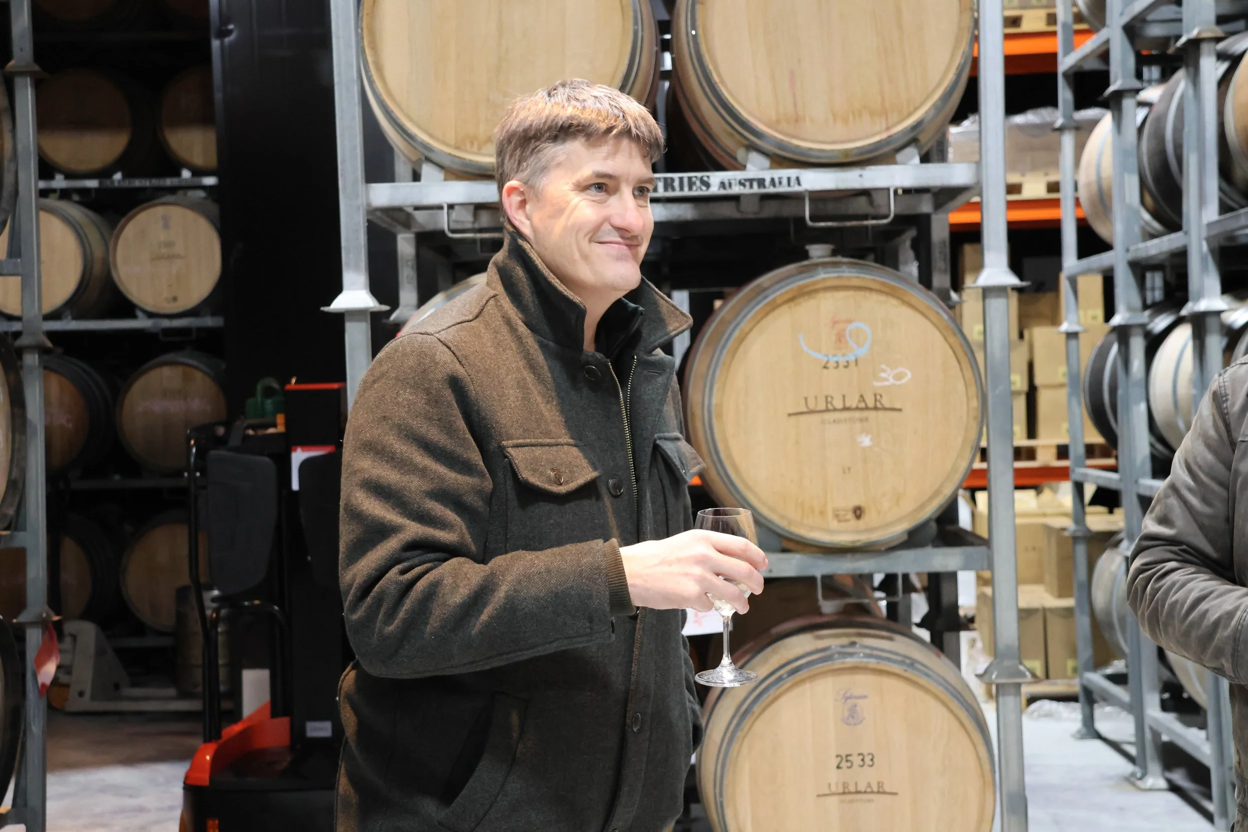 A man holding a wine glass, standing in what appears to be a wine cellar or storage area with large wooden wine barrels in the background.