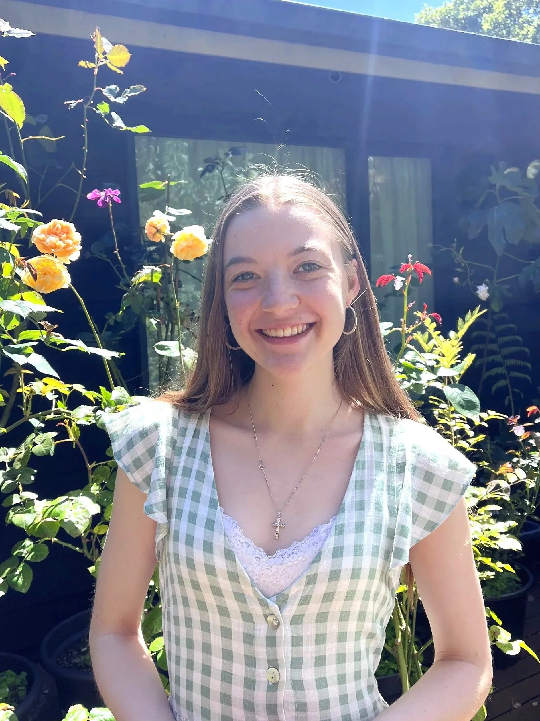 A young woman smiling outdoors in front of colorful flowers and green plants, with sunlight shining.