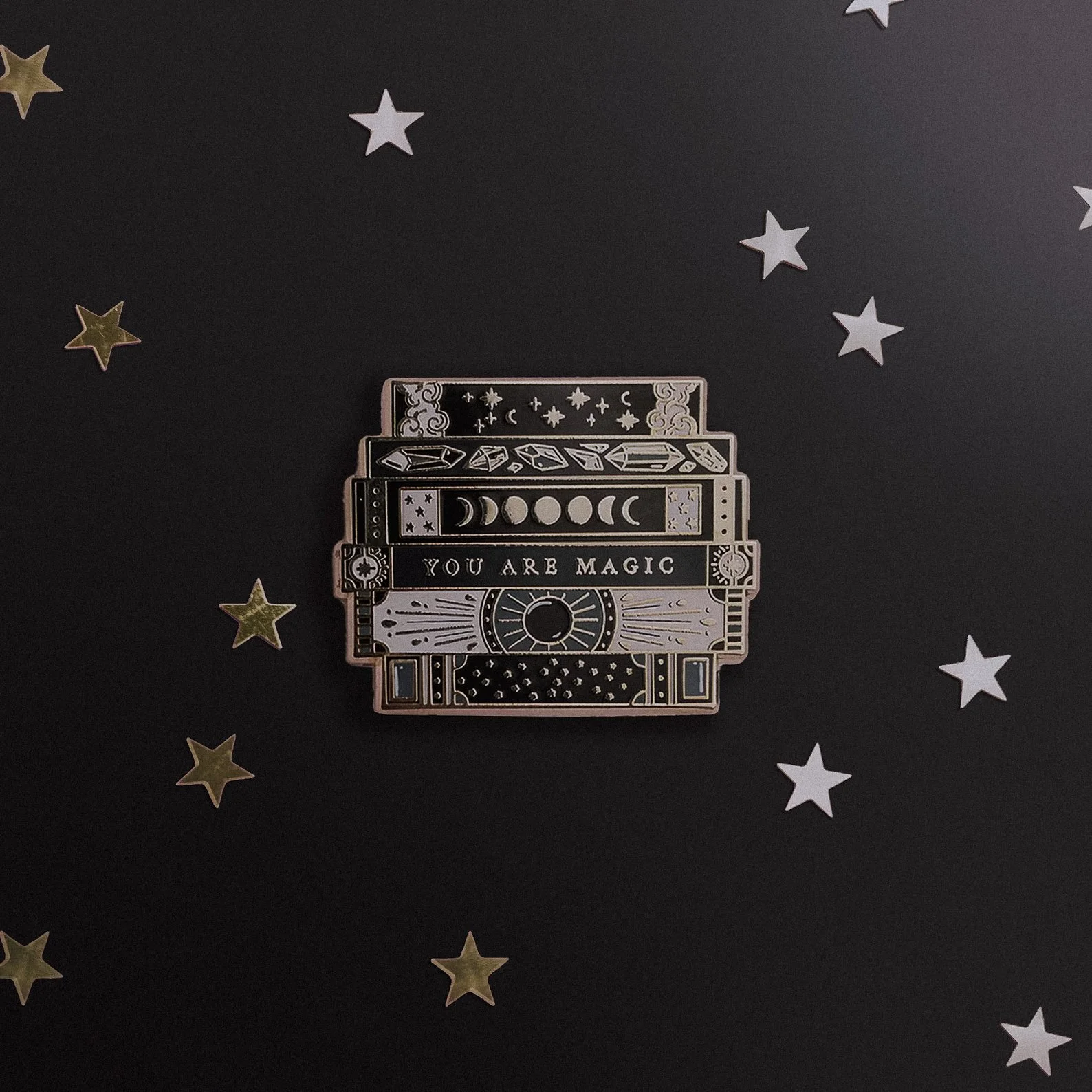 Enamel pin featuring a stack of books adorned with celestial designs in black and gold. Perfect for dark academia enthusiasts, witchy vibes, and bookish fashion statements.