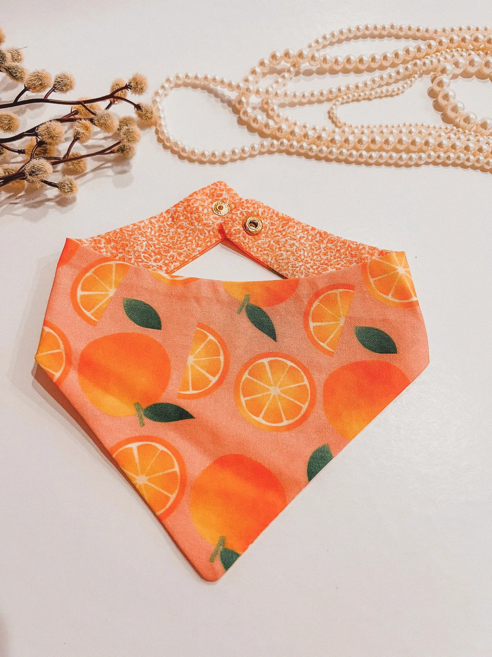 A small dog wearing an orange-themed dog bandana with white polka dots, posing on a white background. The vibrant design features orange slices and citrusy charm.
