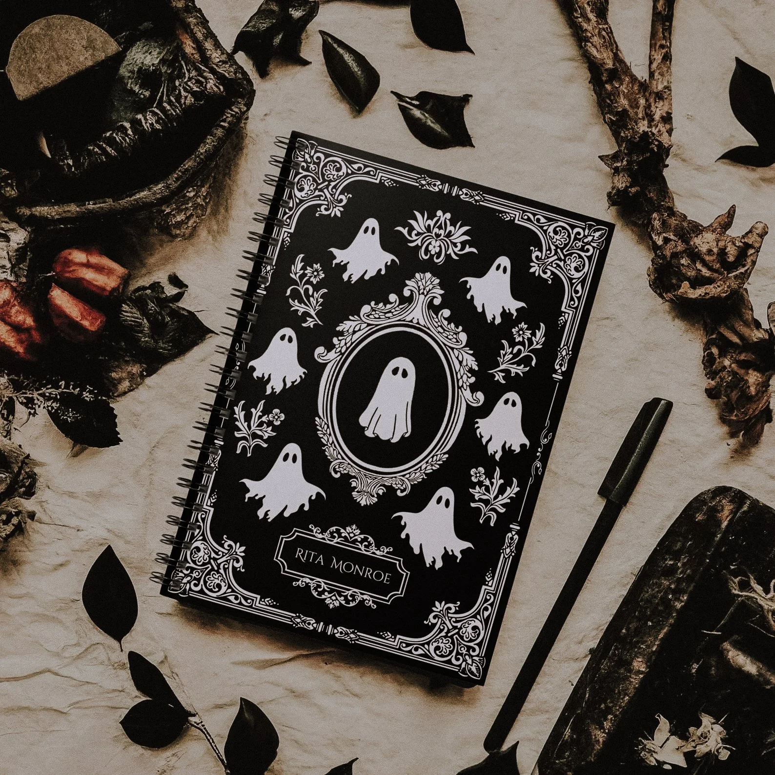 A black and white notebook with ghost illustrations and floral designs on the cover, labeled "Rita Monroe," surrounded by dried leaves, a black pen, and natural decorative objects.