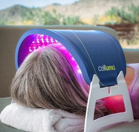 A person with gray hair resting under a blue and purple LED light therapy device labeled 'celluma' on a couch with a mountain view in the background.