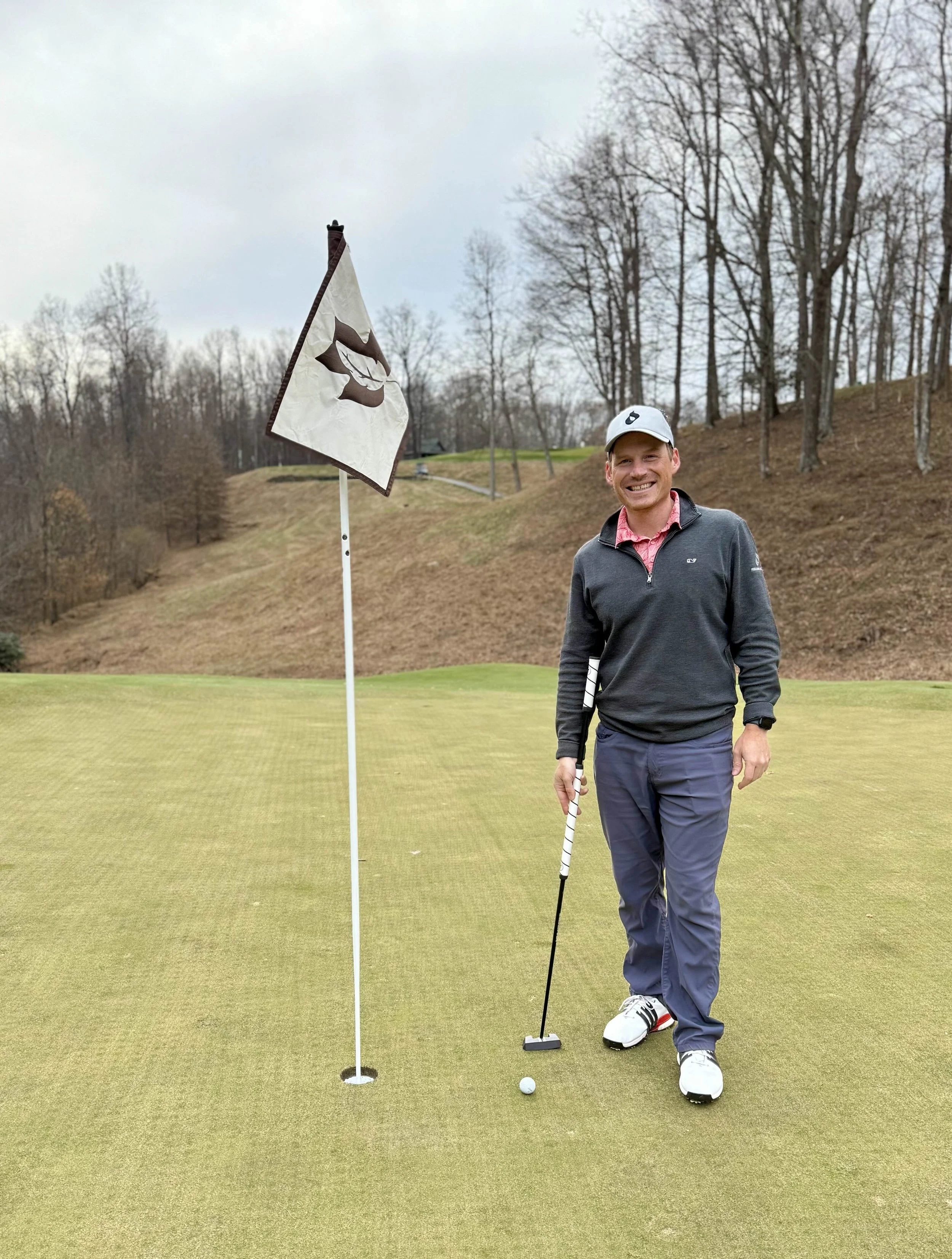 A man standing on a golf green holding a putter, smiling, with a golf flag nearby, surrounded by trees and rolling hills.