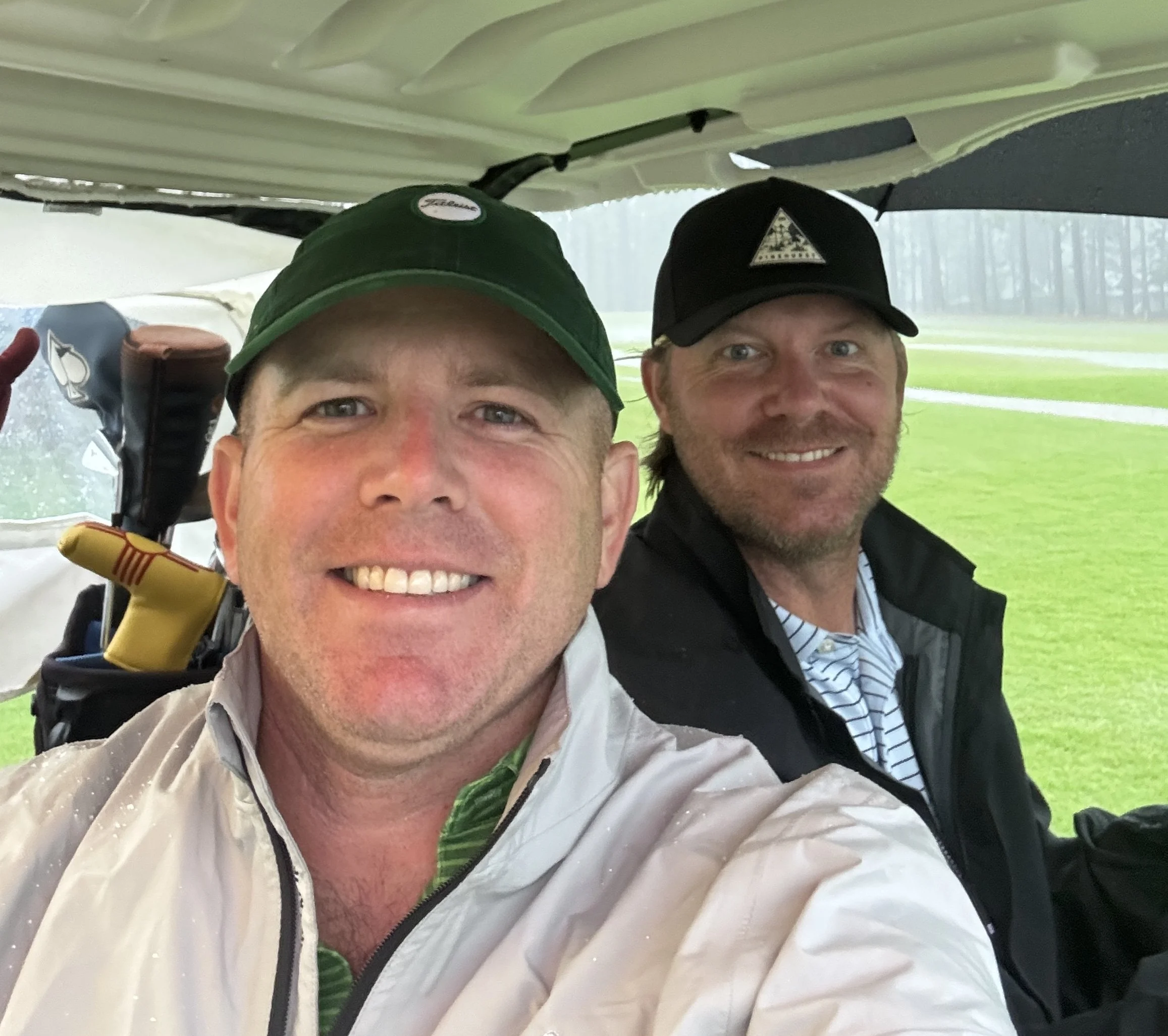 Two smiling men sitting on a golf cart on a rainy day, with a golf bag and clubs in the background, on a golf course with green grass and a misty sky.