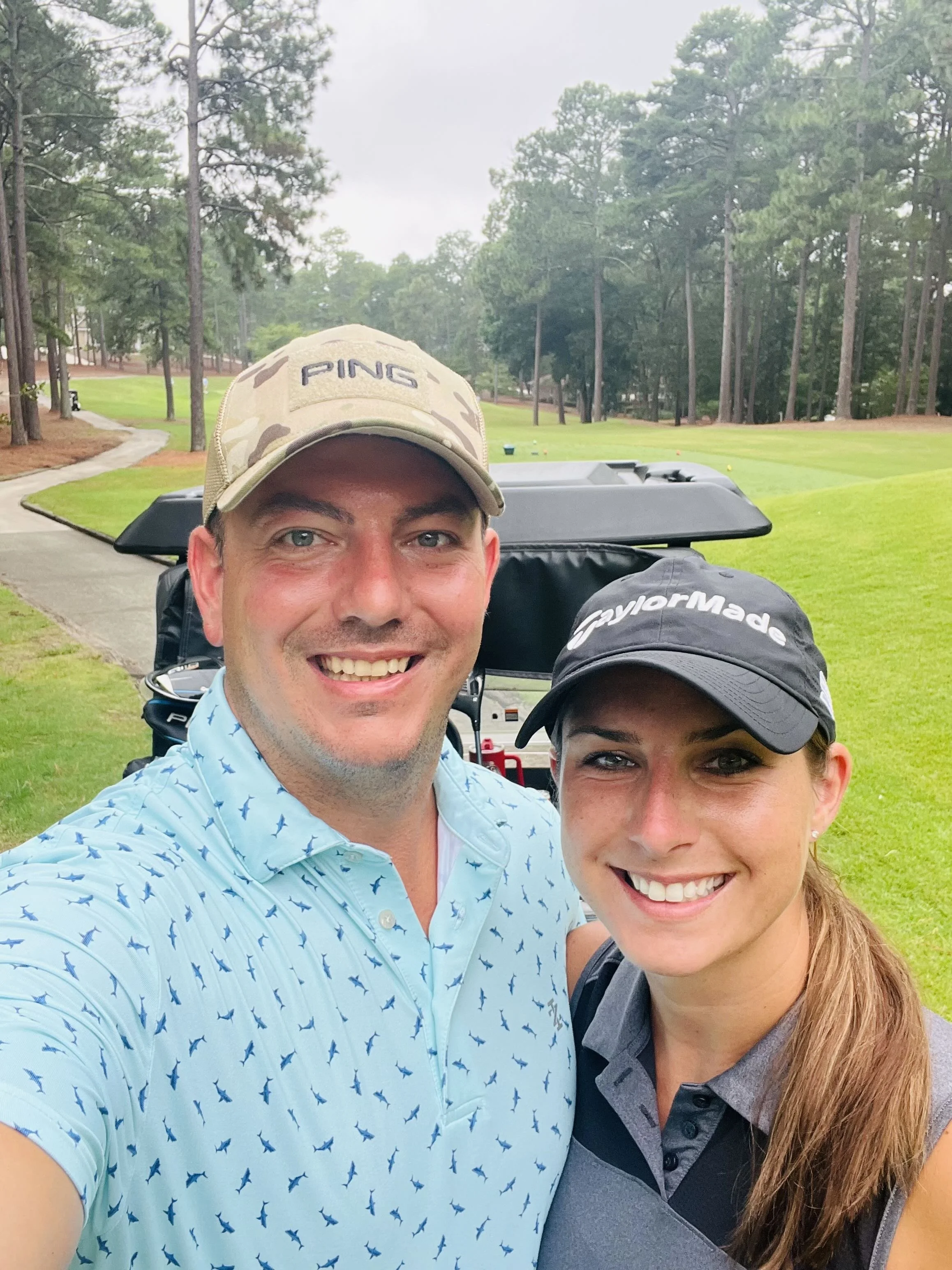A man and woman smiling and taking a selfie on a golf course. The man is wearing a beige camouflage cap and a light blue shirt with small dark patterns. The woman is wearing a black Cap with the TaylorMade logo and a sleeveless golf shirt.