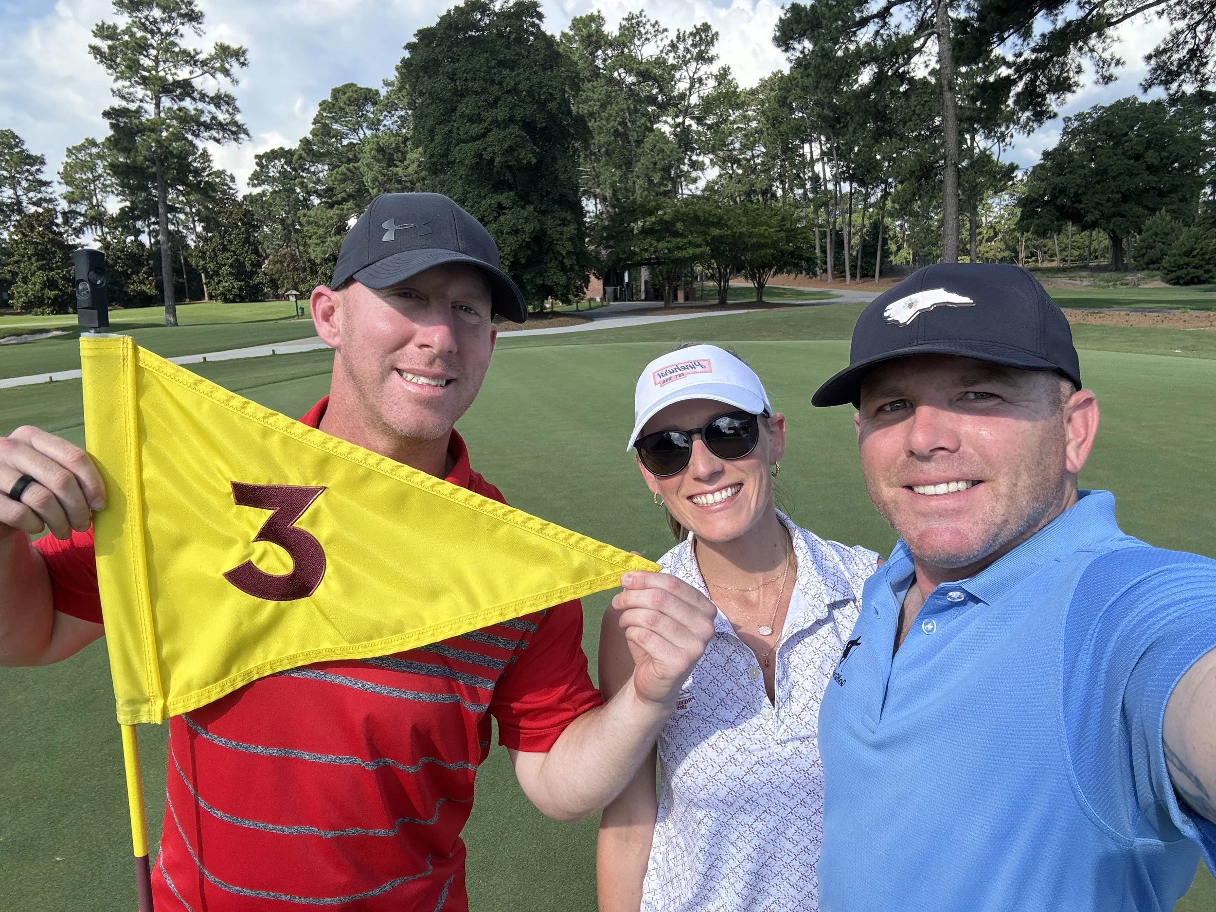 Three people taking a selfie on a golf course with trees and a green in the background. The person on the left is holding a yellow golf flag with the number 3, and everyone is smiling.