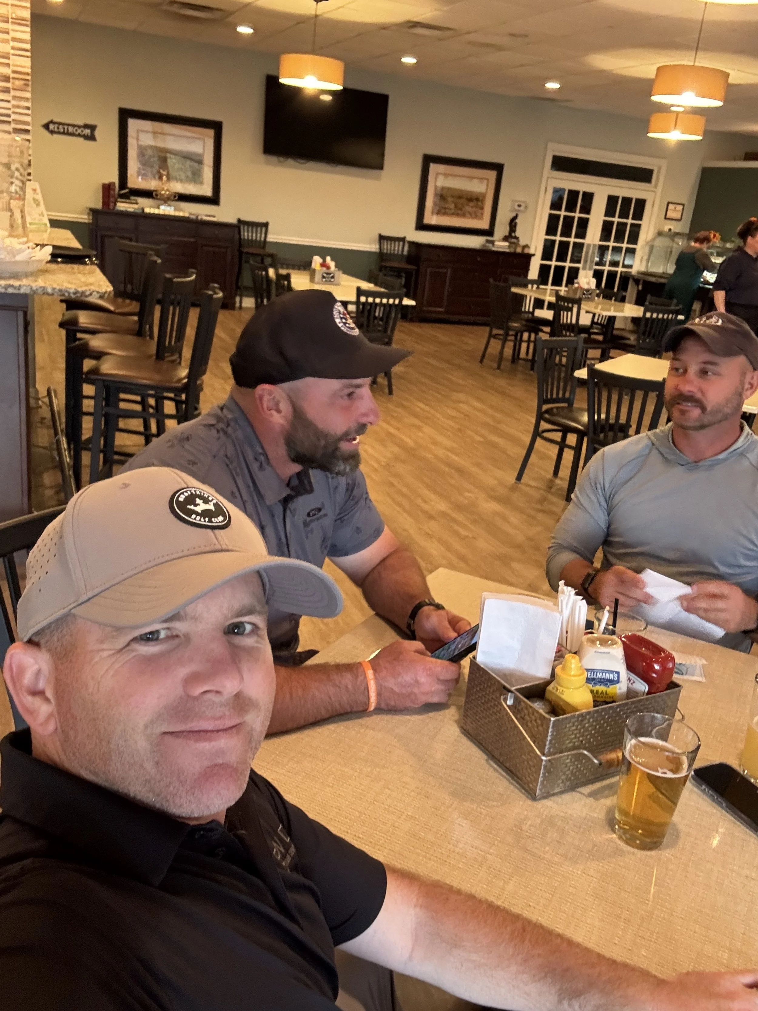 Three men sitting at a restaurant table, with a man taking a selfie, a meat view at the table with condiments, beverages, and utensils, and a restaurant interior with tables, chairs, and a flat-screen TV in the background.