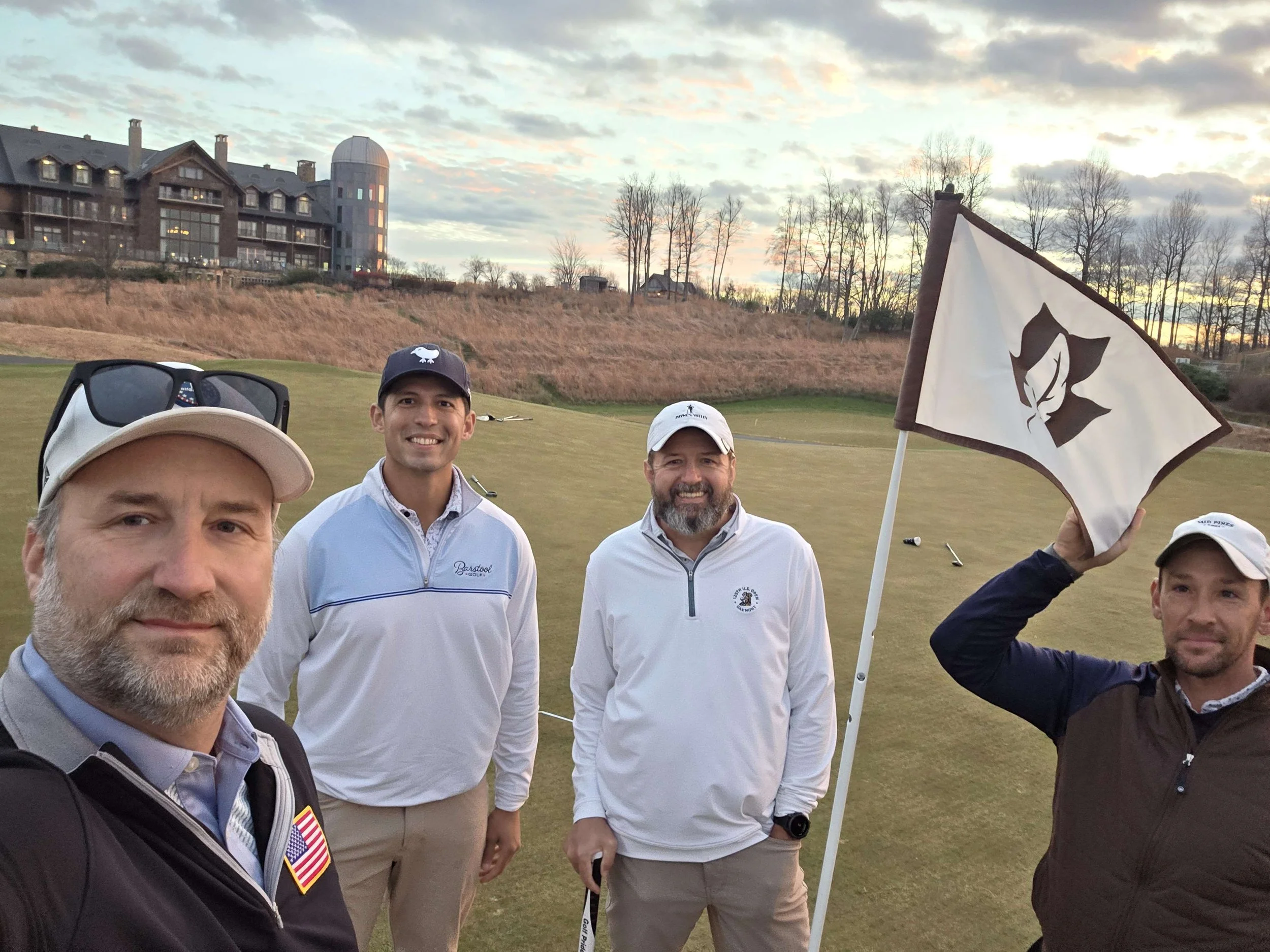 Four men standing on a golf course during sunset, smiling at the camera. One man is holding a flag with a maple leaf logo. The background features a grassy area and a large building with a turret. The men are dressed in golf attire.