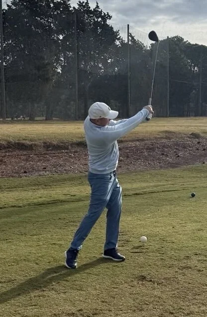 Person in white cap and gray sweatshirt preparing to hit a golf ball on a golf course.