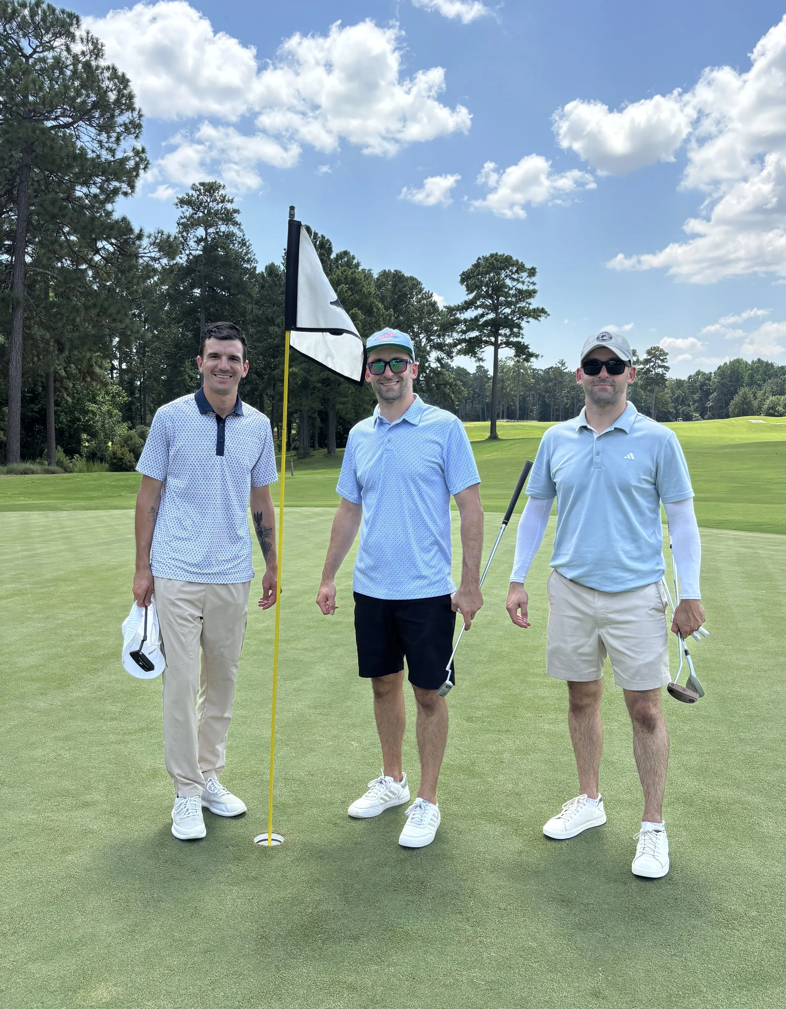 Three men standing on a golf course, smiling, with golf clubs, near a golf hole with a flag, under a partly cloudy sky, surrounded by trees.