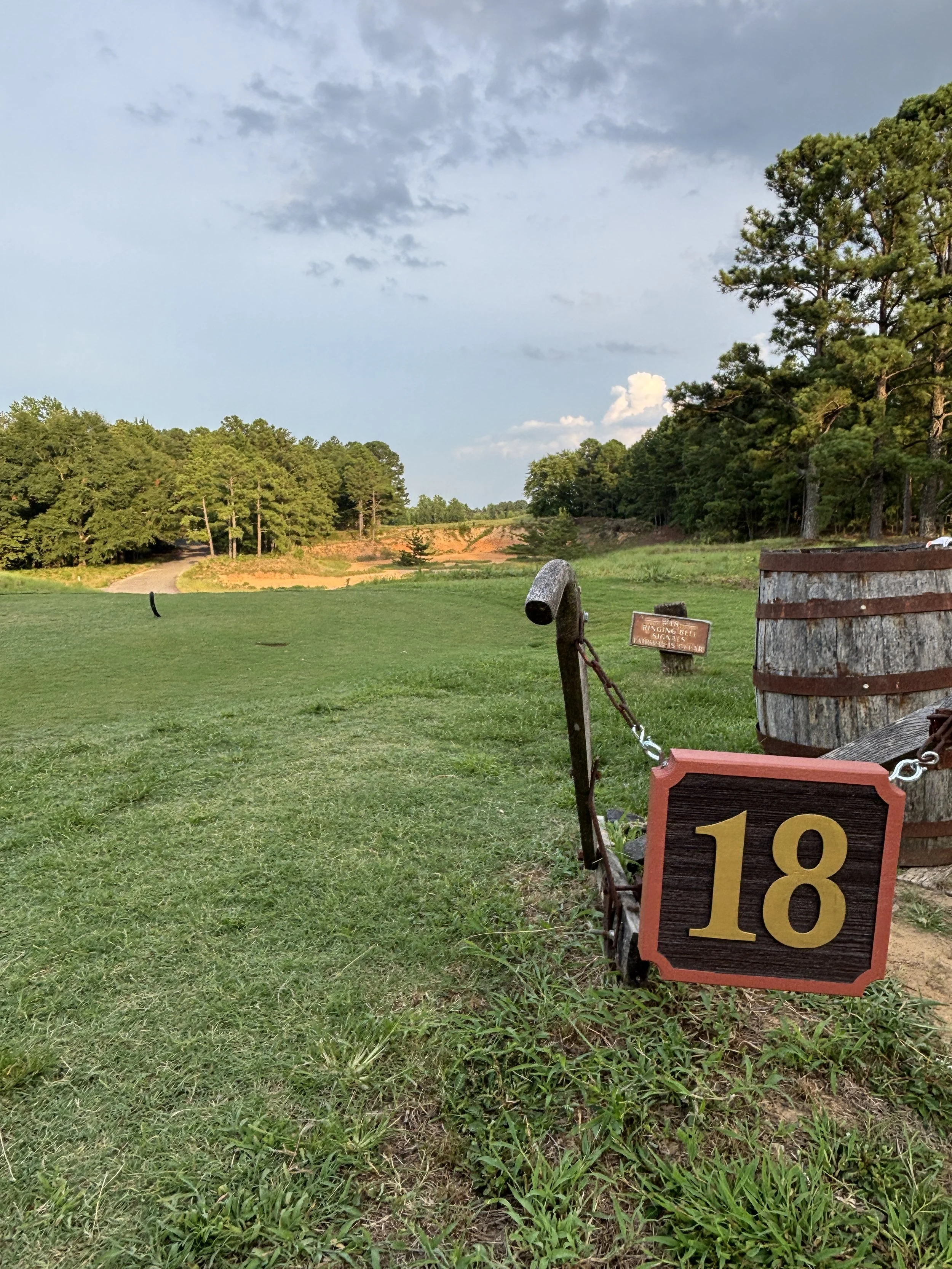 A scenic view of a golf course with a wooden sign displaying the number 18, surrounded by greenery and trees, under a partly cloudy sky.