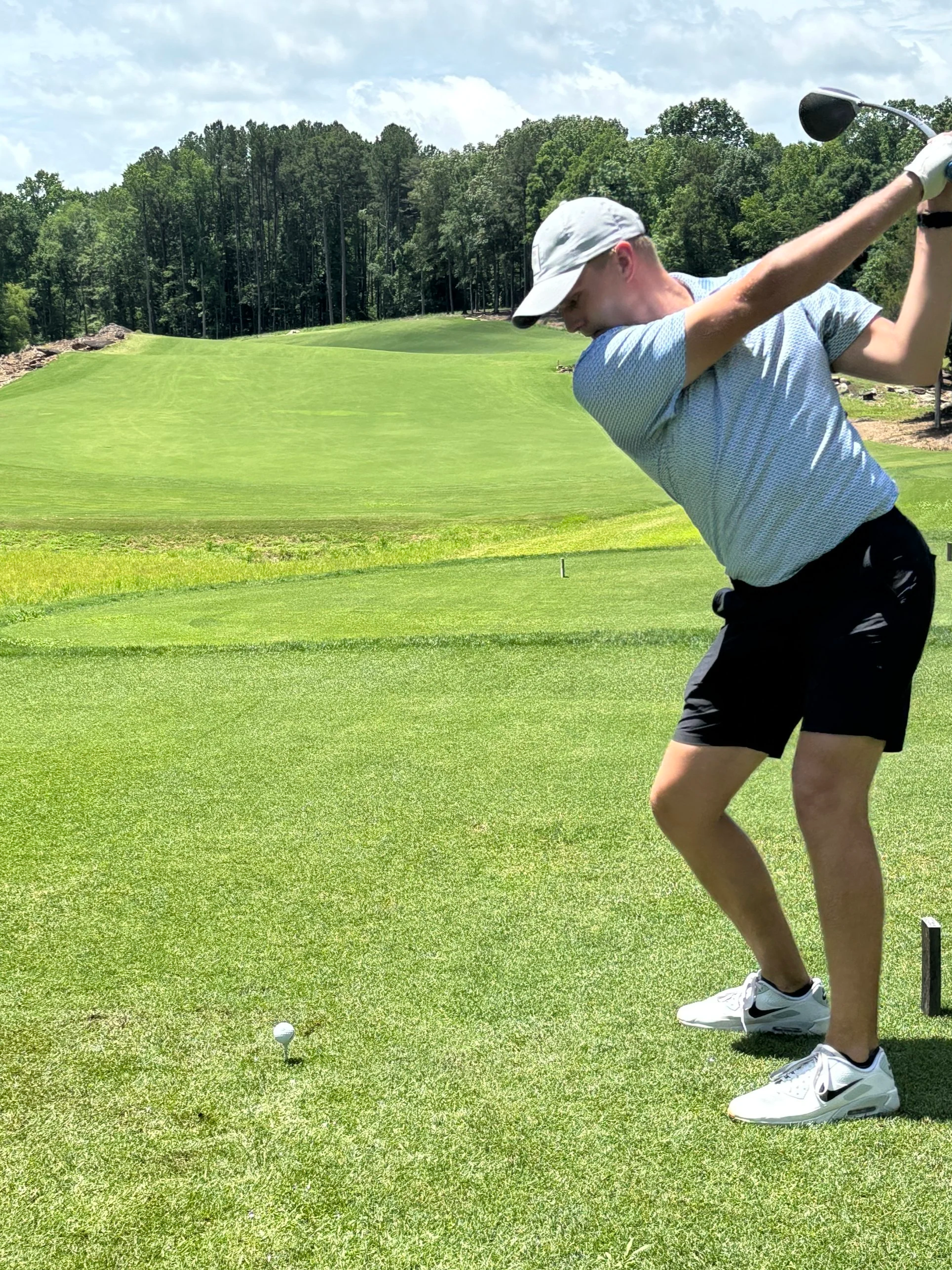 A man dressed in a light-colored cap, gray polo shirt, black shorts, and white Nike sneakers practicing golf on a lush green golf course. He is preparing to hit a golf ball teed up on the grass with a club.