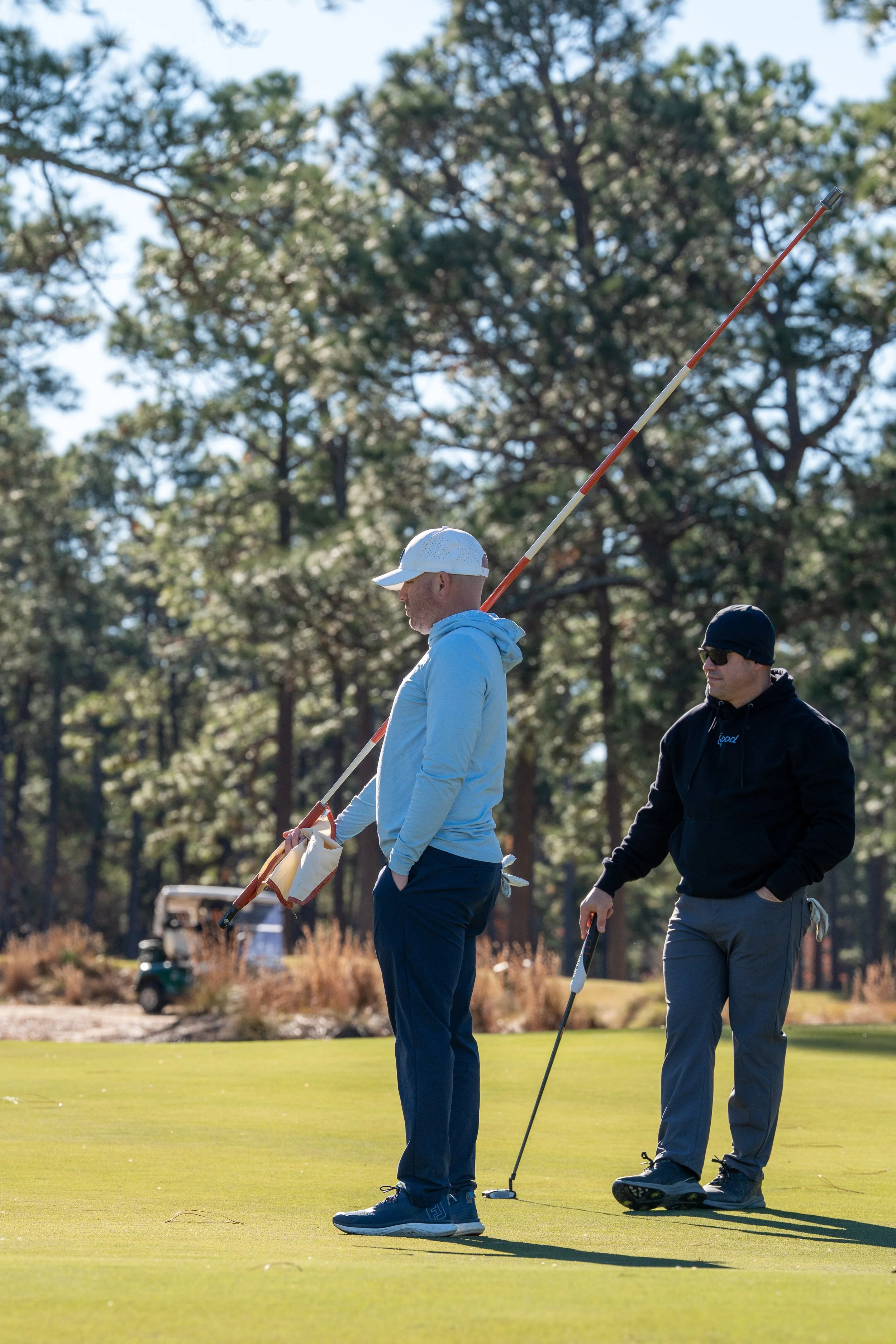 Two men on a golf course, one holding a golf club and the other standing nearby, surrounded by trees and under a clear sky.