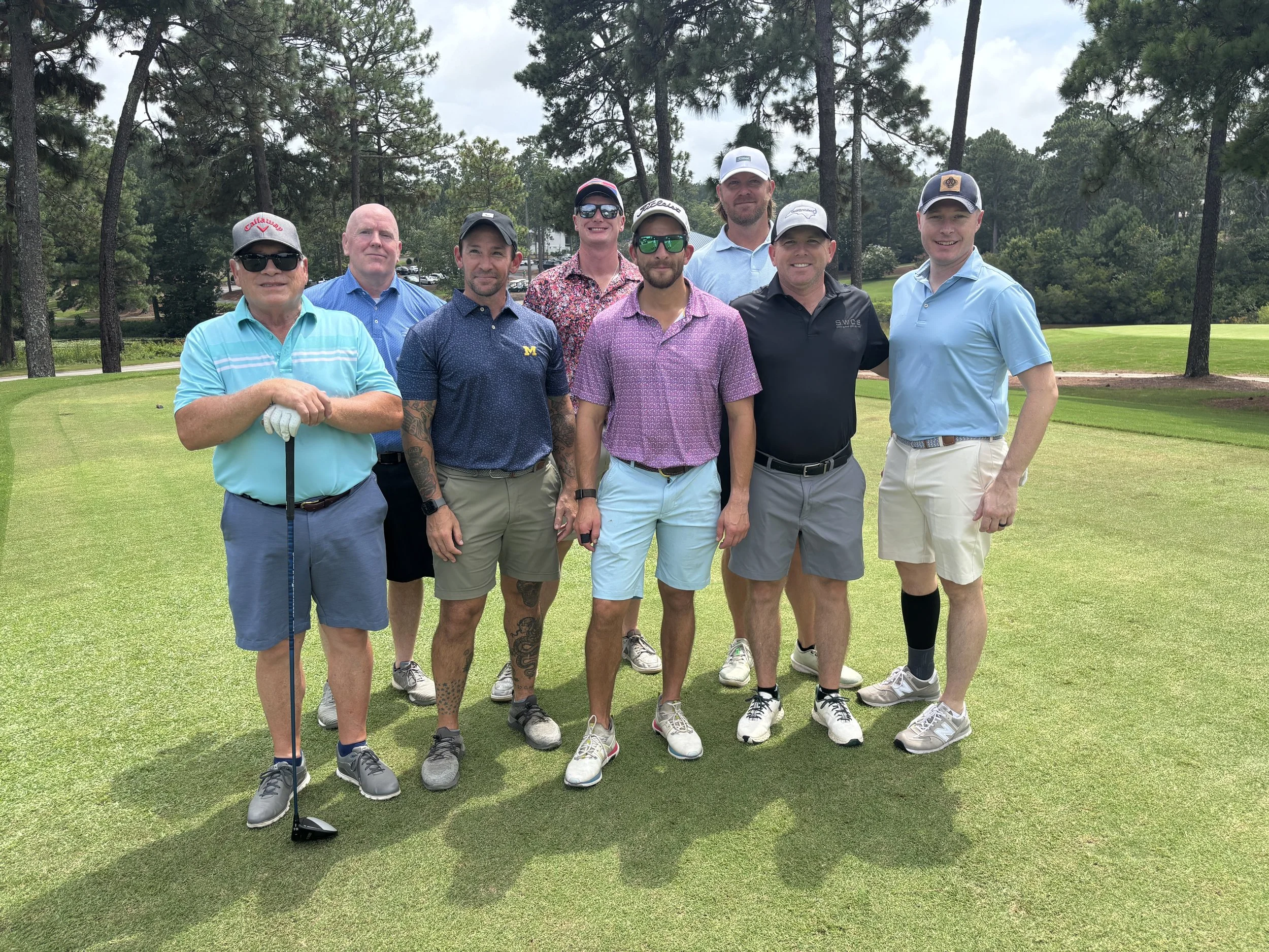 Group of nine men standing on a golf course, dressed in golf attire, with trees in the background.