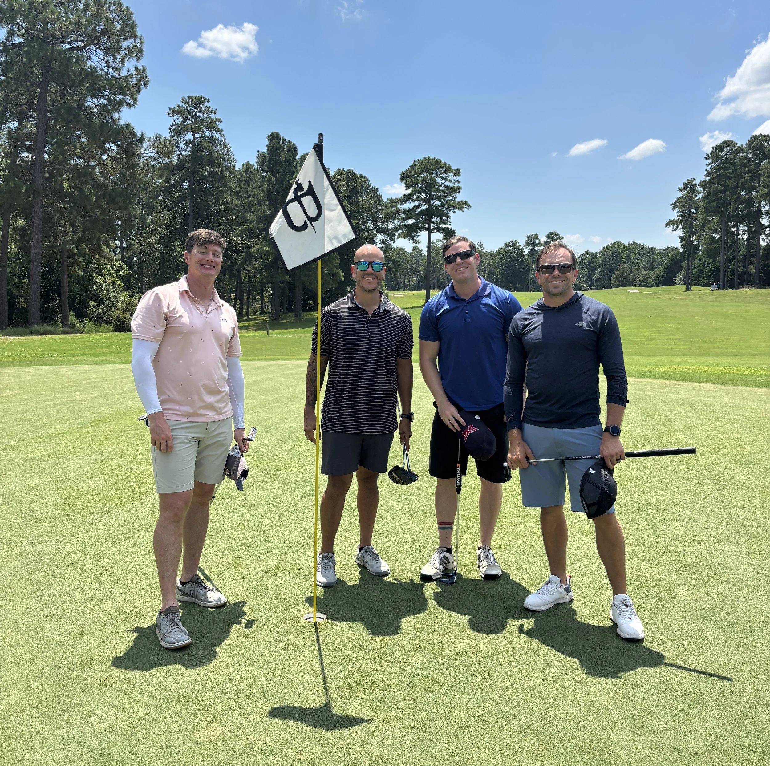 Four men standing on a golf green with a flag on a golf course under a blue sky with some clouds.