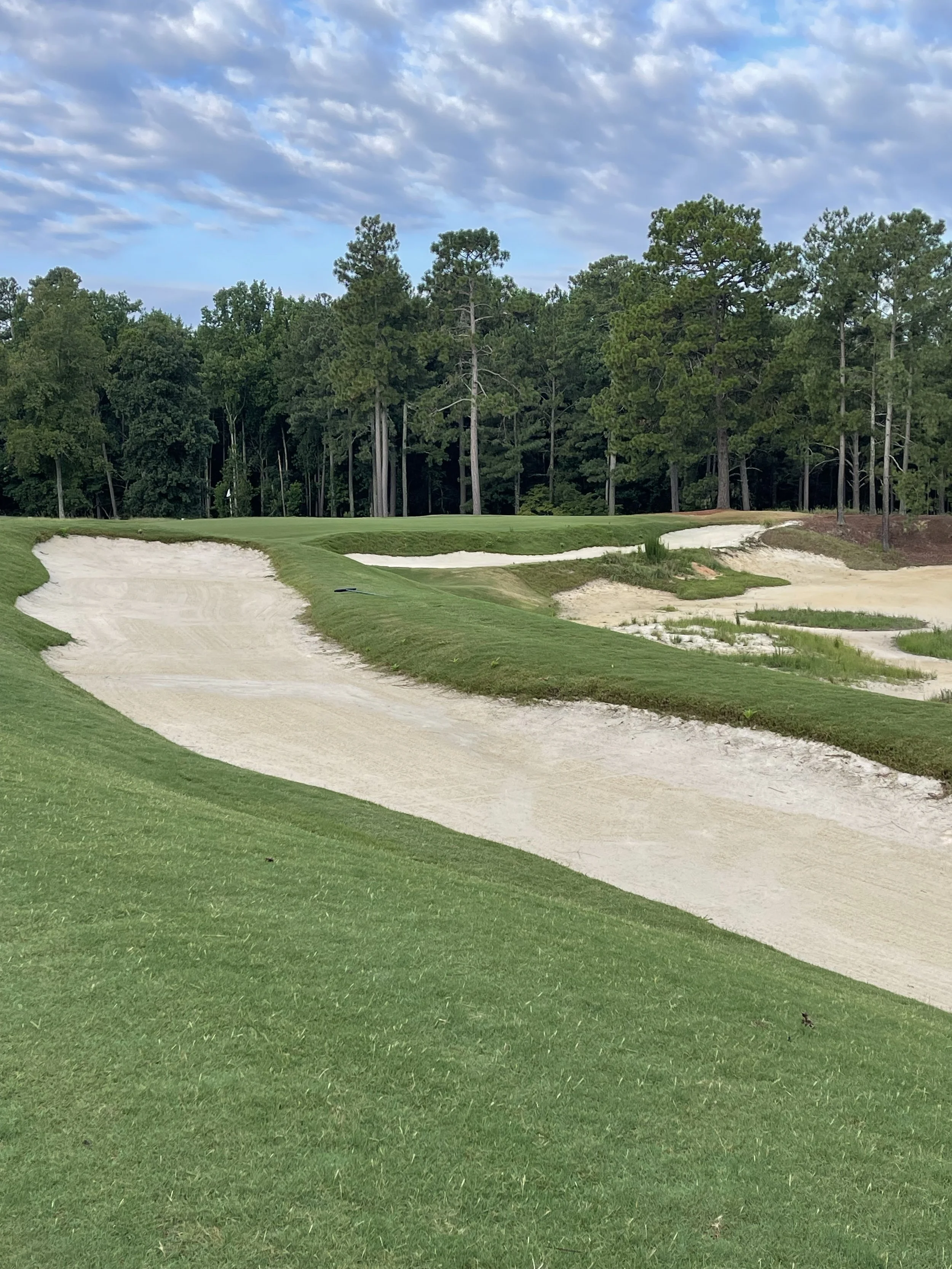 A golf course sand trap with grass edges, set against a backdrop of trees and a partly cloudy sky.