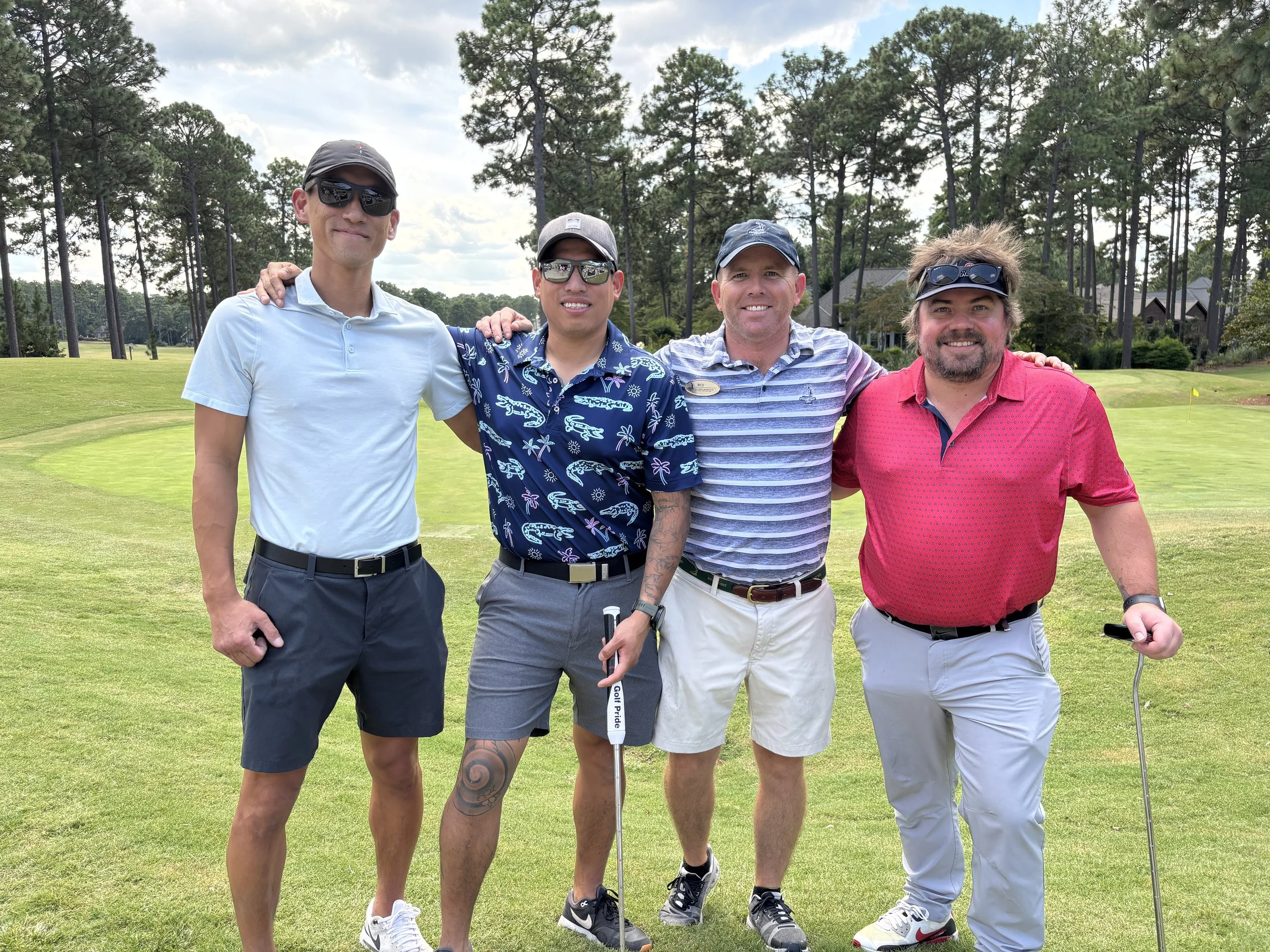 Four men standing together on a golf course, dressed in casual golf attire, with golf clubs and a golf course in the background.