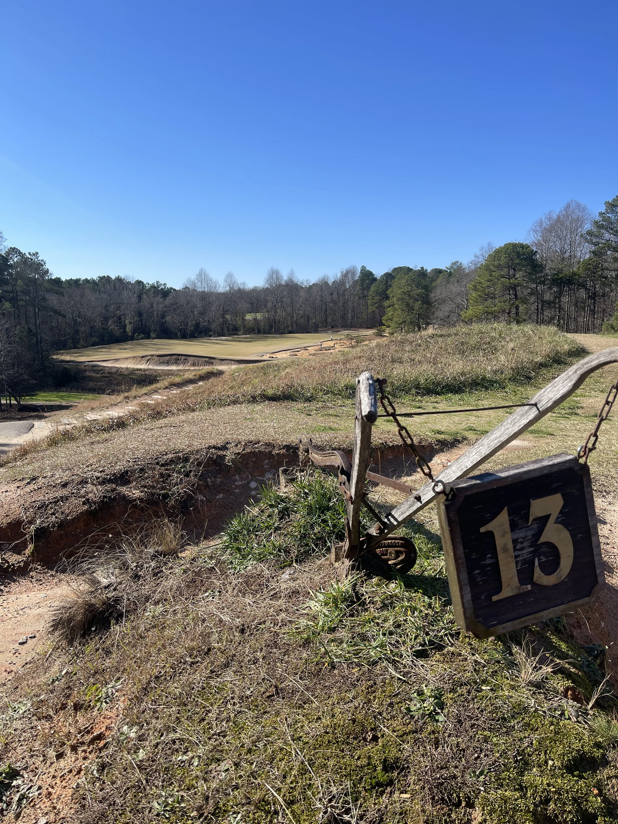 Old unused railroad tie with the number 13, overgrown with grass on a hilly terrain with a golf course and trees in the background under a clear blue sky.