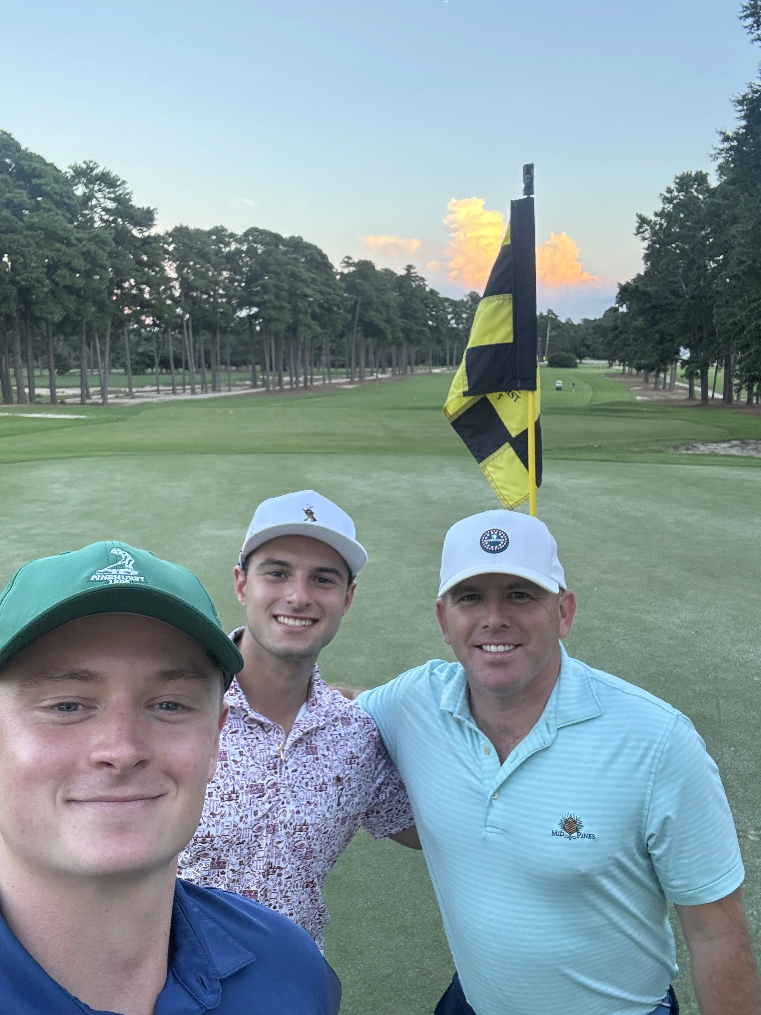 Three men smiling on a golf course, standing near the putting green with a golf flag in the background, surrounded by trees under a partly cloudy sky.