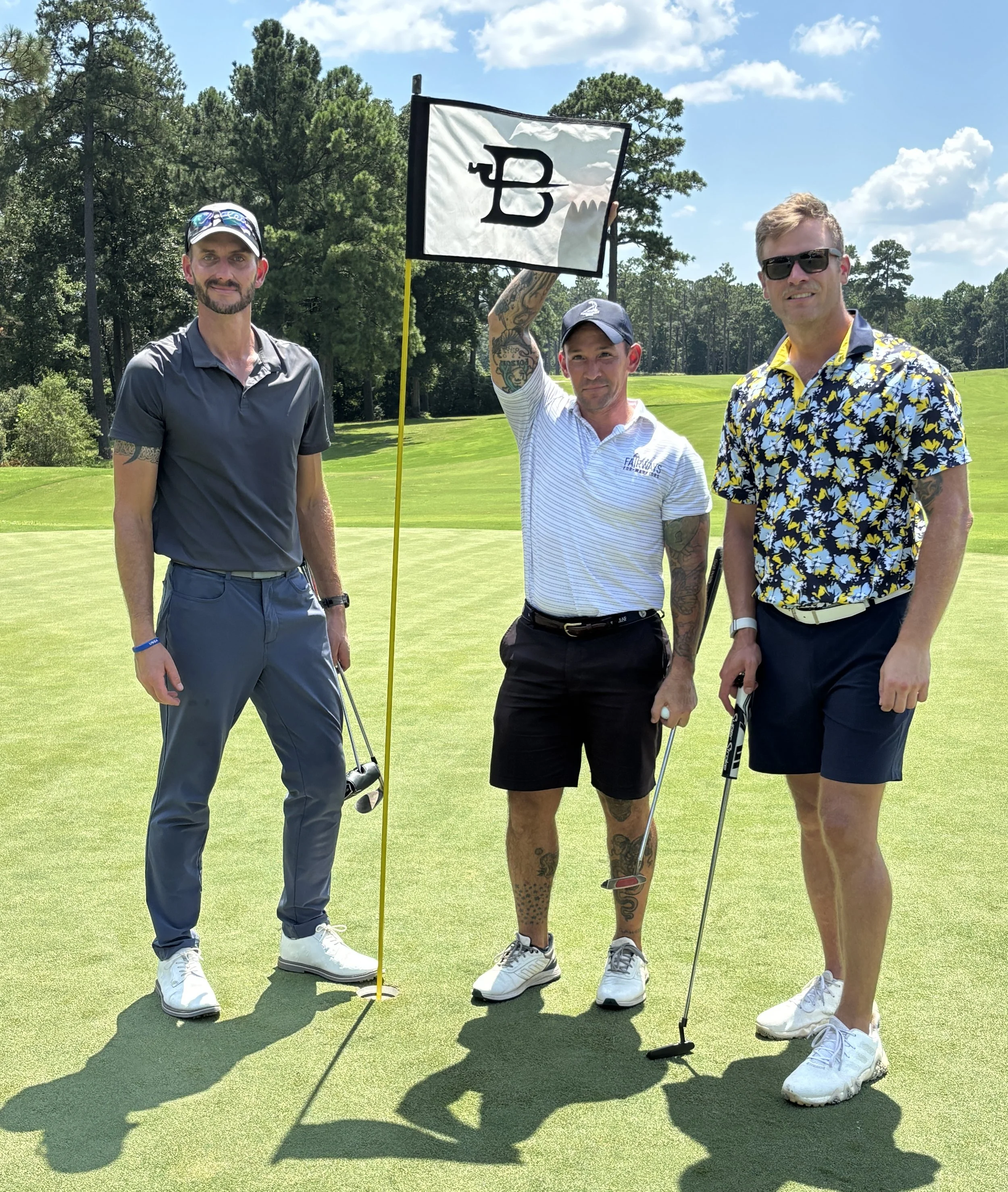 Three men standing on a golf course, one holding a golf flag with a logo, dressed in golf attire, with trees in the background under a blue sky with clouds.