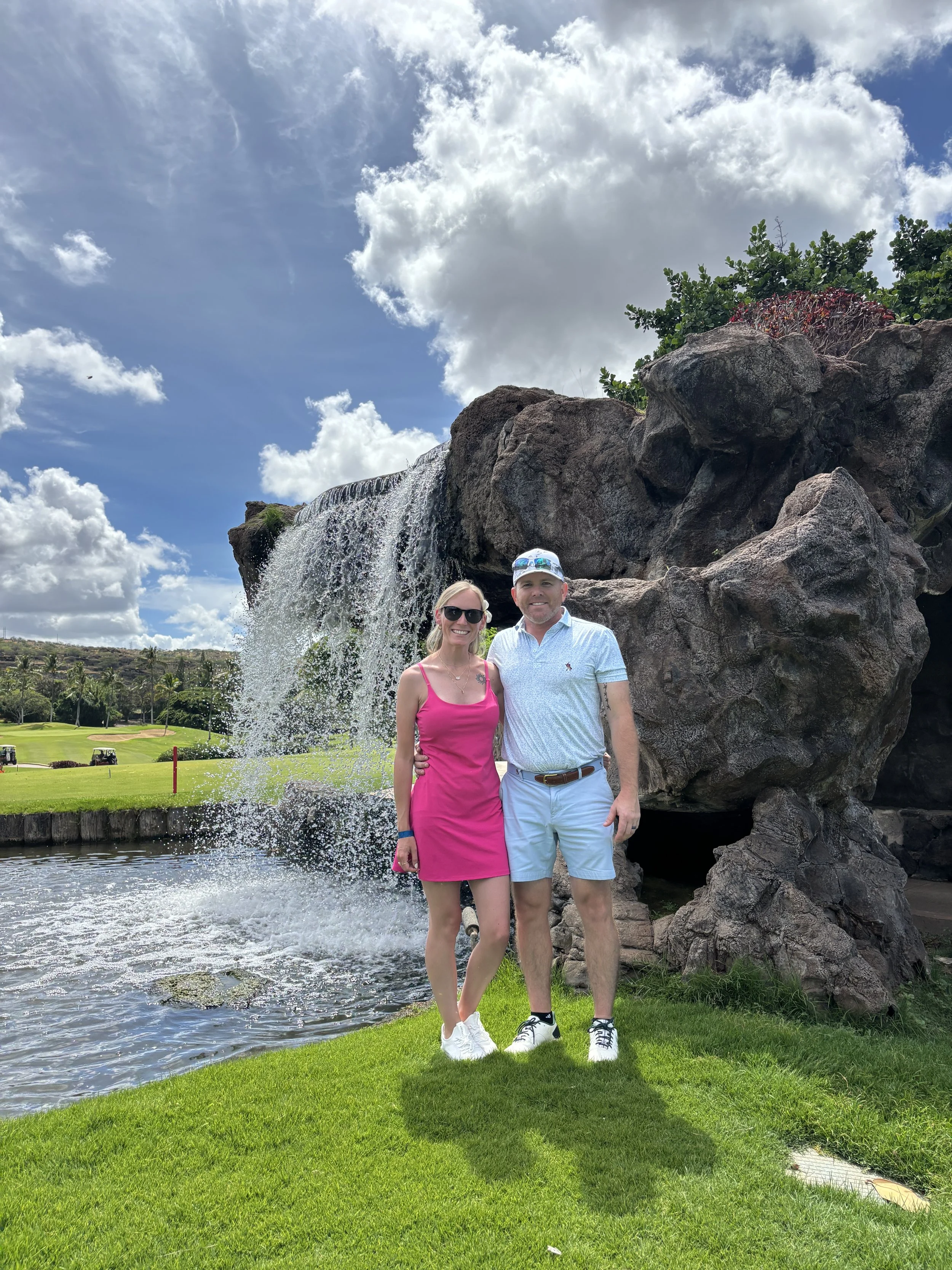 A couple standing on a golf course near a waterfall with rocks, green grass, and a partly cloudy blue sky.