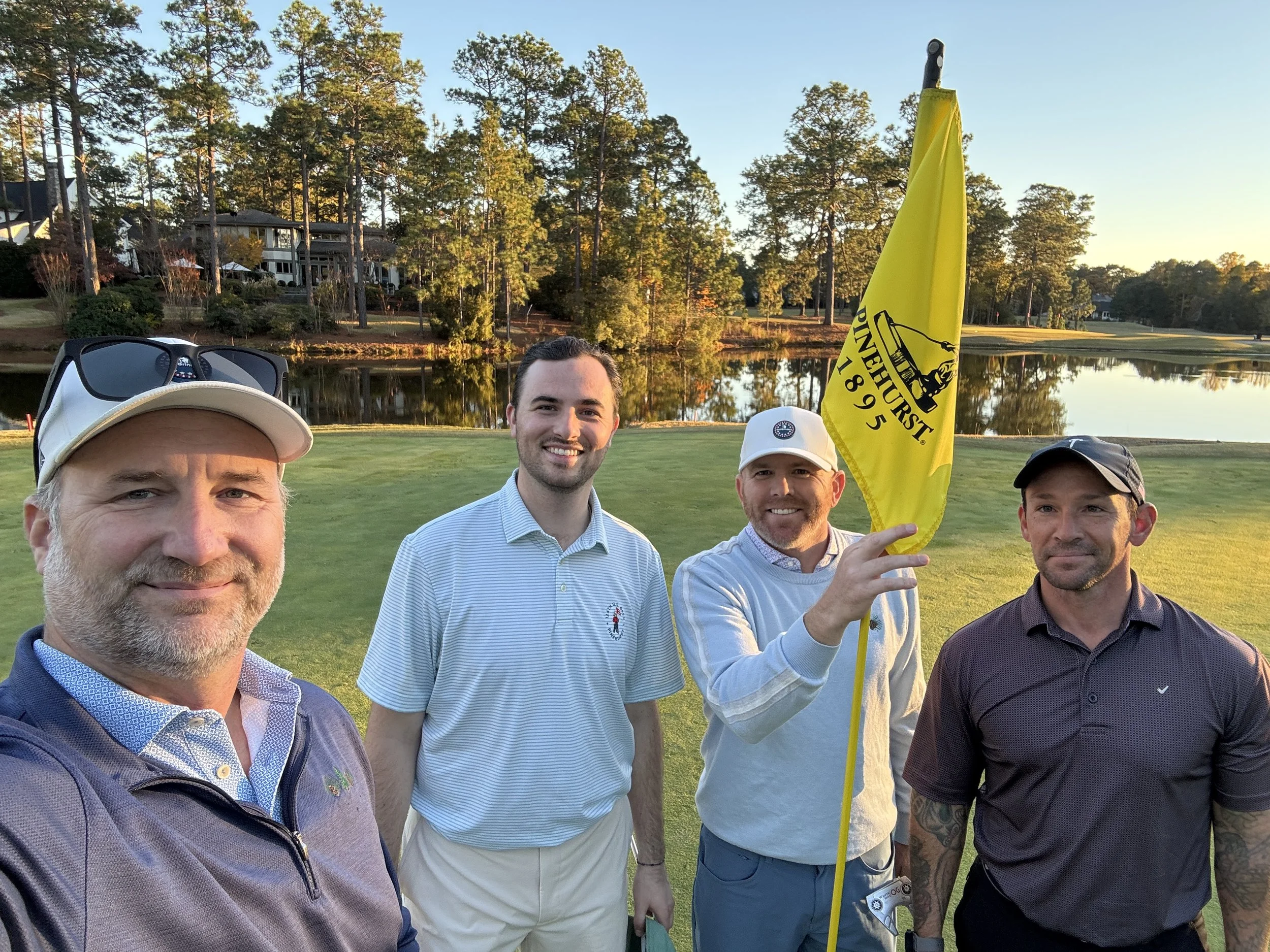 Four men taking a selfie on a golf course during sunset, with one man holding a yellow flag with a black horse logo and the words "Horse Inn 1895", a water hazard, trees, and houses in the background.